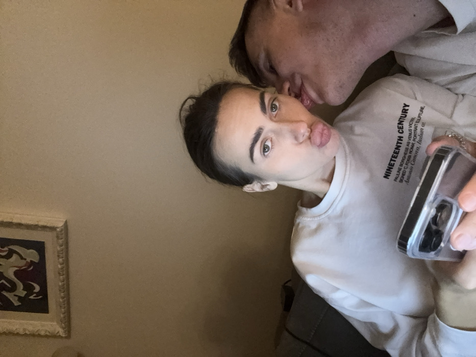 The image depicts two individuals indoors. The specific location within Venice, Italy, or even the city itself, cannot be identified from the visual evidence presented.
A female individual, appearing to be an adult, is positioned centrally, facing the camera. She has dark hair styled back and light-colored eyes, displaying a puckered lip expression. She wears a light-colored, long-sleeved crew-neck sweatshirt with the text "NINETEENTH CENTURY PAULINE BORGHESE AS VENUS VICTRIX SIGNED C. FOSSI ROMA PORTRAIT SCULPTURE Antonio Canova, Italian art" printed across the chest. Her right hand holds a smartphone encased in a clear cover, revealing multiple camera lenses on its back. A silver-colored chain or bracelet is visible on her wrist or hand.
To her right, a male individual, also appearing to be an adult, is partially visible, showing the right side of his face and neck with short, dark hair. He is leaning towards the female's face, with his tongue extended and making contact with her right cheek/jaw area.
The background consists of a plain, light beige or off-white wall. In the upper left quadrant, a portion of a framed artwork is visible; it features abstract or stylized shapes in dark red and lighter tones, set within an ornate, light-colored frame. The lighting within the scene is warm and ambient.