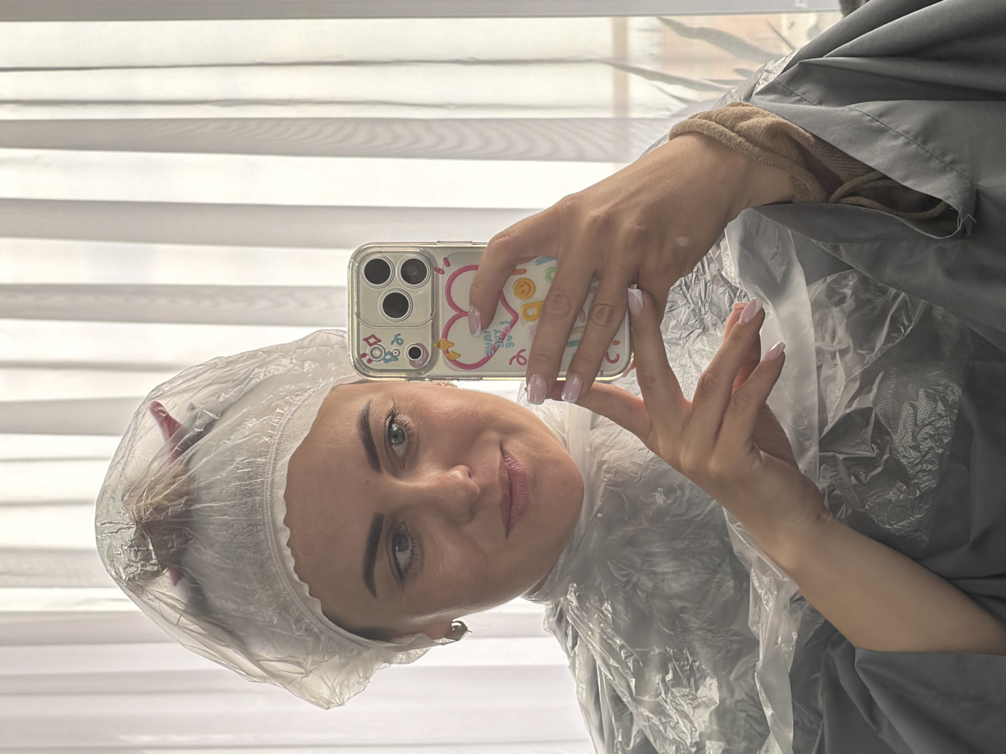 The image shows a woman taking a selfie. She is wearing a plastic shower cap and a plastic protective cape, suggesting she is at a hair salon or undergoing a hair treatment. She is holding a smartphone with a decorated case, likely using it to capture the image. The background features a set of horizontal blinds. There is no indication of the city Votkinsk, Russia in the image.