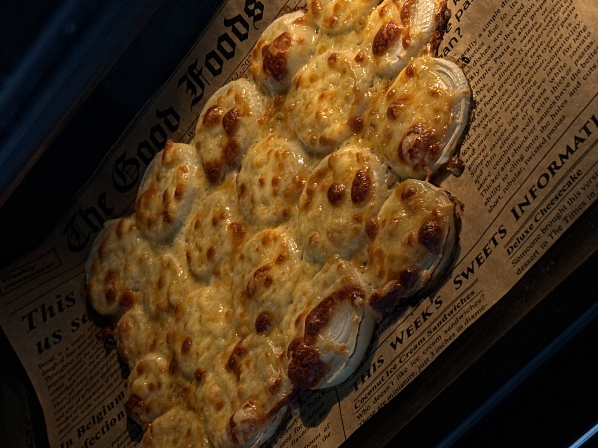 A close-up, overhead shot showcases a tray of baked cheese-topped onion rings, presented on a sheet of newspaper. The food is golden brown and appears to be a savory appetizer or side dish. The newspaper serves as a rustic backdrop, with visible text and headlines, suggesting a casual dining context. The lighting is dramatic, with strong highlights and shadows, emphasizing the texture of the melted cheese and the crisp edges of the onion rings. The scene is static, with no visible people or actions, creating a still-life composition focused entirely on the food and its presentation. The specific location of Lyubertsy, Russia, is not ascertainable from the visual information provided in the image.