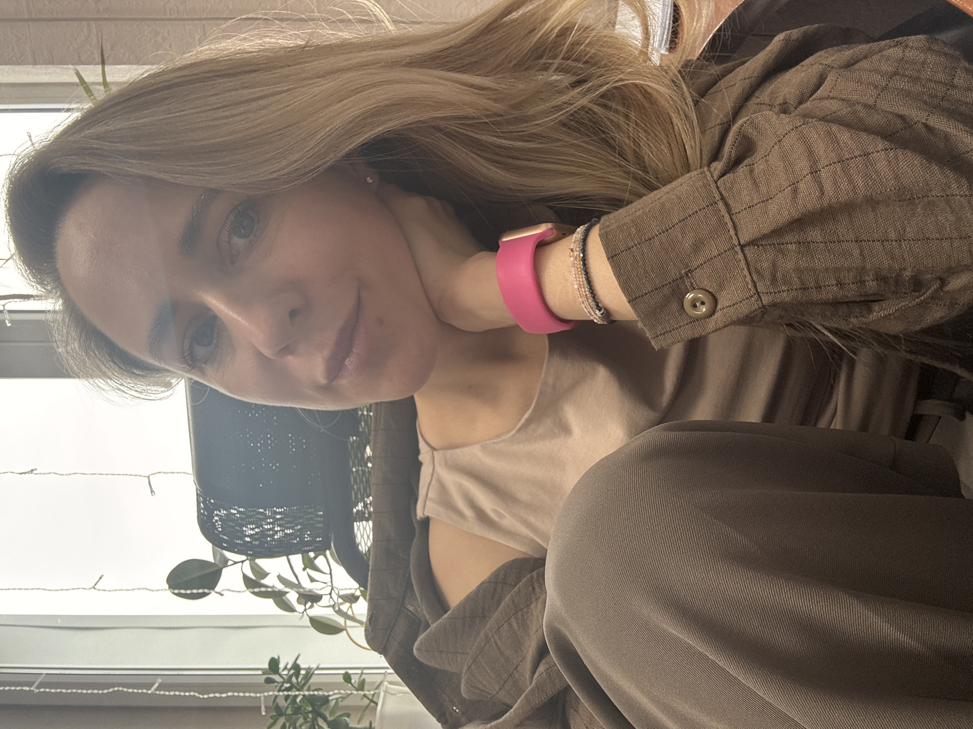 A woman with long blonde hair is posing for a selfie. She is wearing a brown button-down shirt over a beige tank top. She is also wearing brown pants. On her wrist, she has a pink watch and a bracelet. She is holding her hand to her neck. In the background, there is a window with plants and fairy lights. Based on the user request, the location is Chaikovsky, Russia.