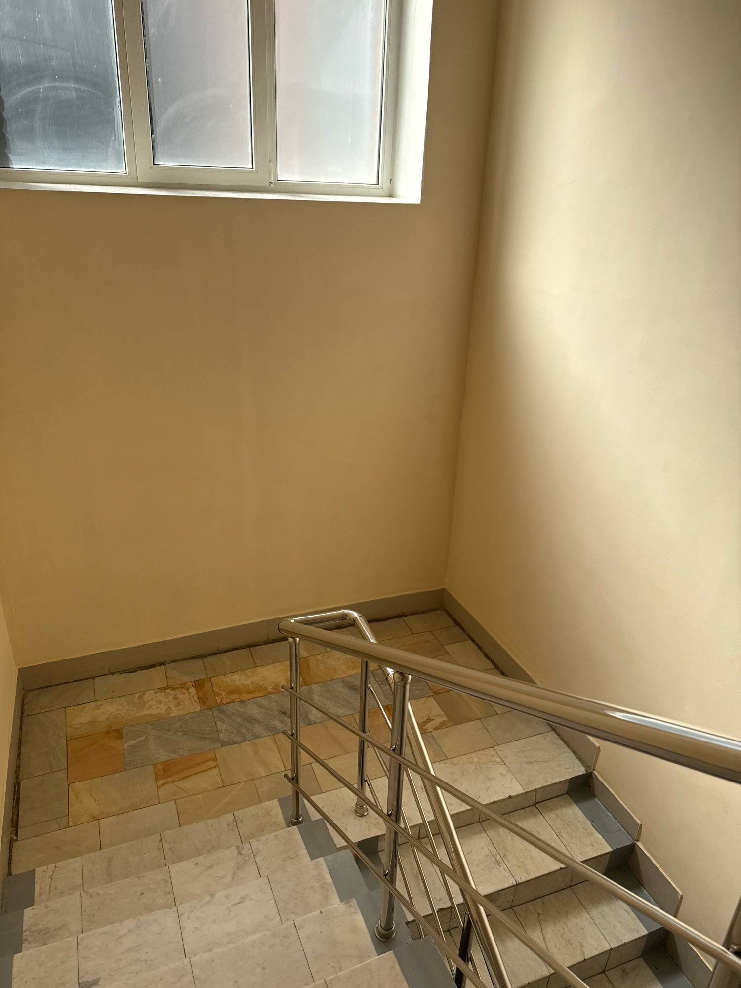 An indoor shot captures a staircase with a metal railing, leading down from a landing. The landing is tiled with a mix of light-colored marble and stone tiles in shades of beige, brown, and grey. The walls are painted a pale yellow, and a band of grey tiling runs along the base of the wall. Above the landing, a horizontal window with frosted panes is visible. The stairs themselves are also tiled with light-colored marble. The scene is lit by natural light, casting shadows on the walls and stairs. There are no people present.