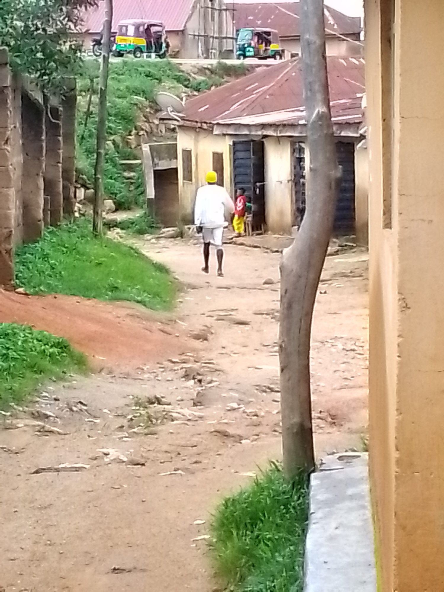 An outdoor, eye-level shot captures a person wearing a white robe and a yellow cap walking away from the camera down a dirt path in a village. To the left of the path, green grass and a reddish-brown dirt embankment are visible. Further down the path, several buildings with corrugated metal roofs and shuttered windows line the sides. In the background, a tri-colored auto-rickshaw, yellow, green, and red, is parked on an incline. A thin, vertical tree trunk stands prominently in the foreground to the right, obscuring a portion of the right side of the image, where a light-colored building wall is visible.