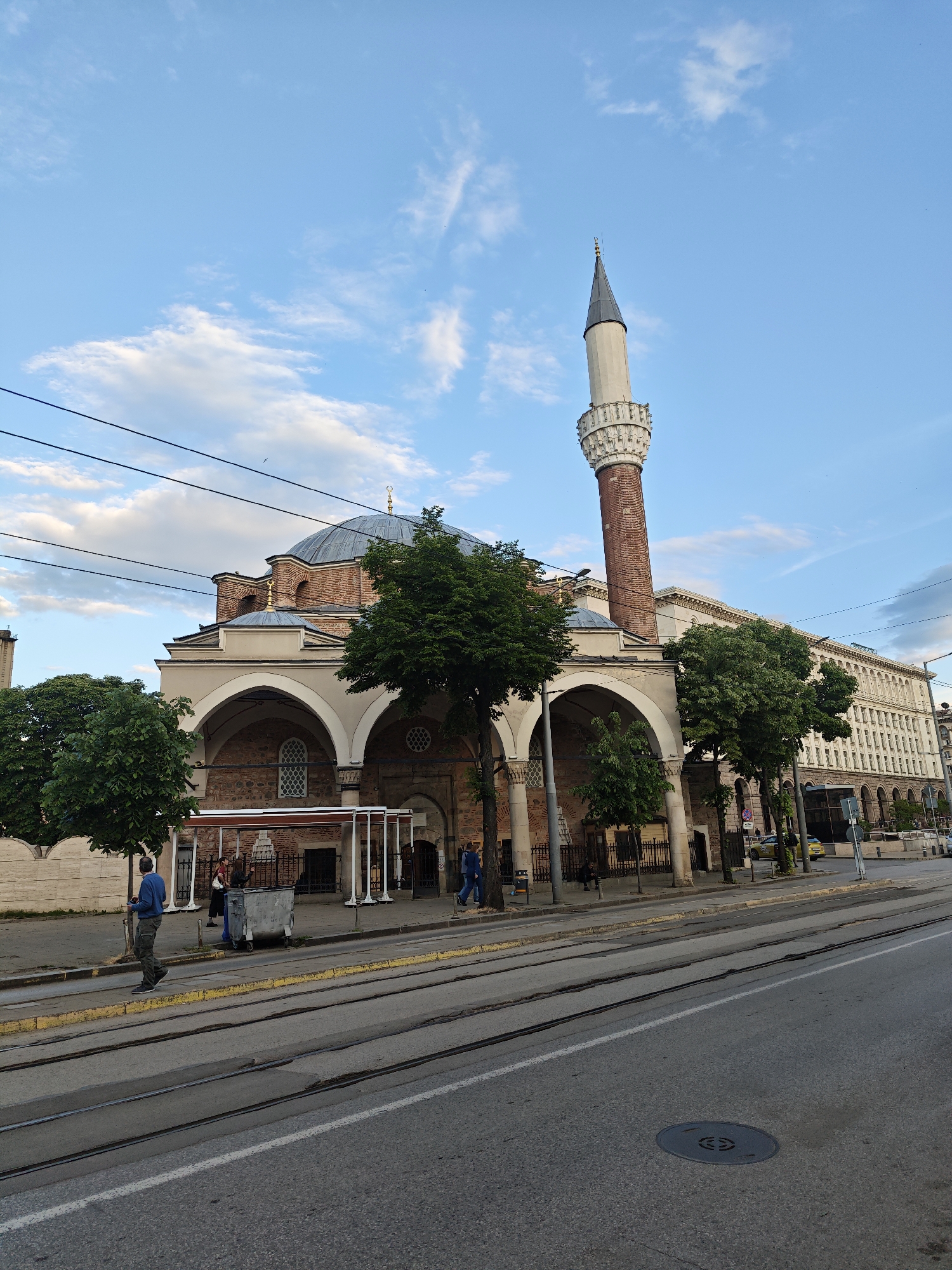 The image captures a view of the Banya Bashi Mosque in Sofia, Bulgaria. The mosque features a brick and stone facade with arched windows and a prominent minaret. A dome-shaped roof tops the main structure. In front of the mosque, there are tram tracks running along the street. A group of people stand near a bus stop. Trees line the sidewalk, adding a touch of greenery. In the background, a large building is visible, indicating the urban setting. The sky is partially cloudy. A man in a blue shirt and khaki pants is walking on the sidewalk. A metal trash bin stands near the bus stop. Overhead power lines are visible against the sky.
