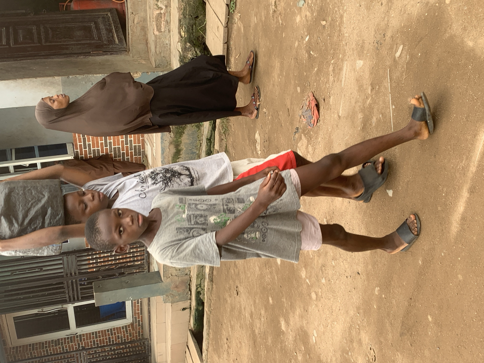 In Shagamu, Nigeria, on a dirt path, a young boy wearing a grey t-shirt and pink shorts walks towards the viewer. Behind him, another person, whose face is partially obscured, carries a large dark bag on their head. To the right, a woman wearing a brown hijab and a black skirt walks away from the viewer. The background consists of a building with brick walls and metal gates, and a wooden structure. Scattered on the ground are a few discarded items, including what appears to be a torn shoe or sandal.