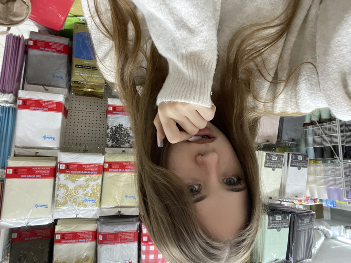 A woman wearing a white sweater poses for the camera in a store aisle. She's biting her nail with a piercing in her nose. Behind her is a variety of packaged tablecloths and other plastic-wrapped goods. The rows of merchandise create a colorful backdrop, with shades of red, white, blue, and gold visible. The image is oriented in a rotated or upside-down position.