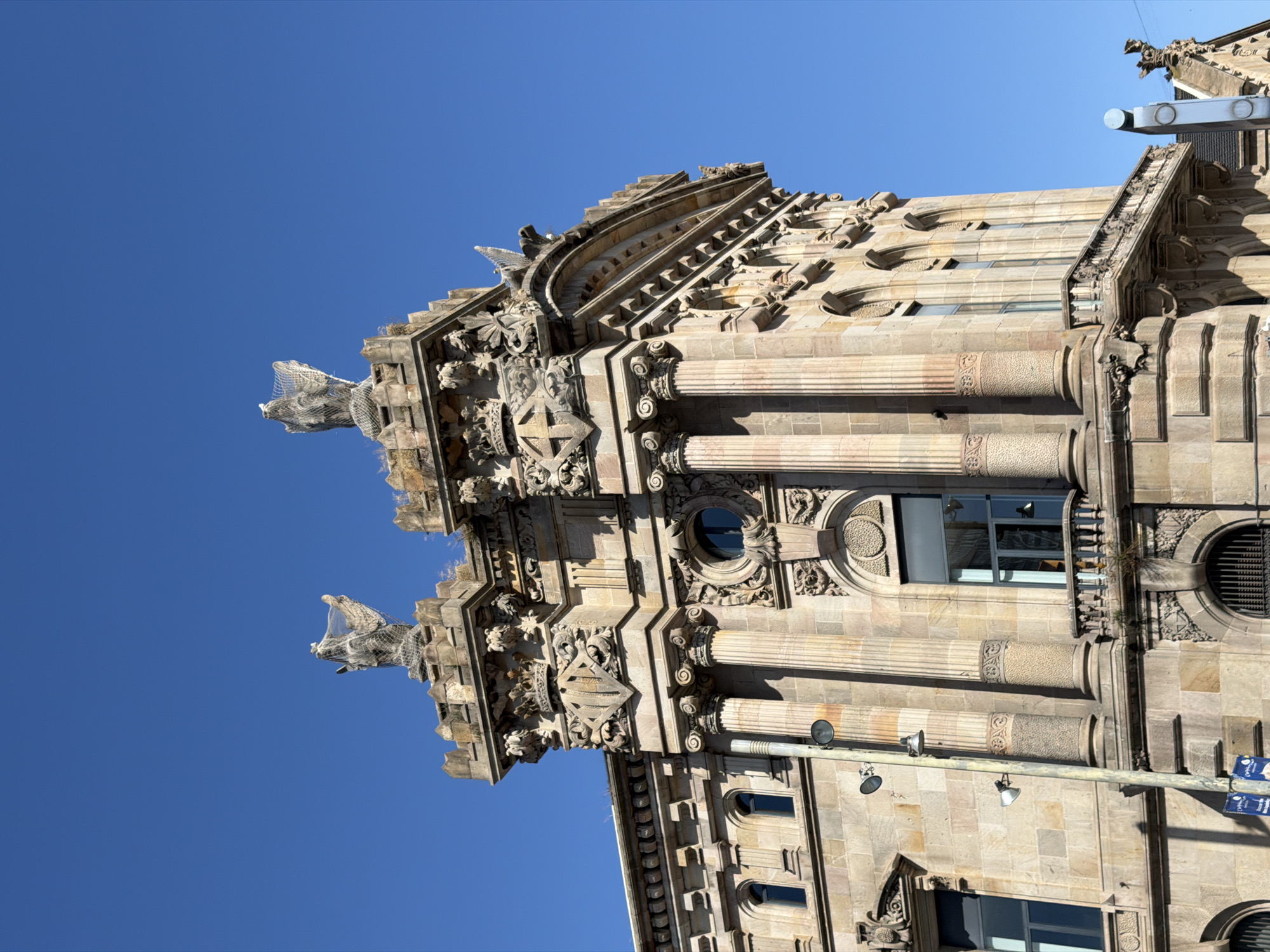 This media file captures a low-angle view of an ornate building facade against a clear, bright blue sky, indicating a sunny day. The building, made of light-colored stone, features intricate architectural details, including columns, sculptures, and decorative carvings. Two winged figures or gargoyles are prominent, one on the upper left with its head covered in netting, and another on the lower left, also partially obscured by netting. The style of the architecture suggests it is a historic building. The scene is outdoors, and the lighting suggests it is daytime. There are no people or specific activities depicted; the focus is solely on the building's architecture. Location cues include street lights and what appear to be street signs, one of which has text that is partially visible but unreadable. The overall impression is one of historical grandeur and detailed craftsmanship.