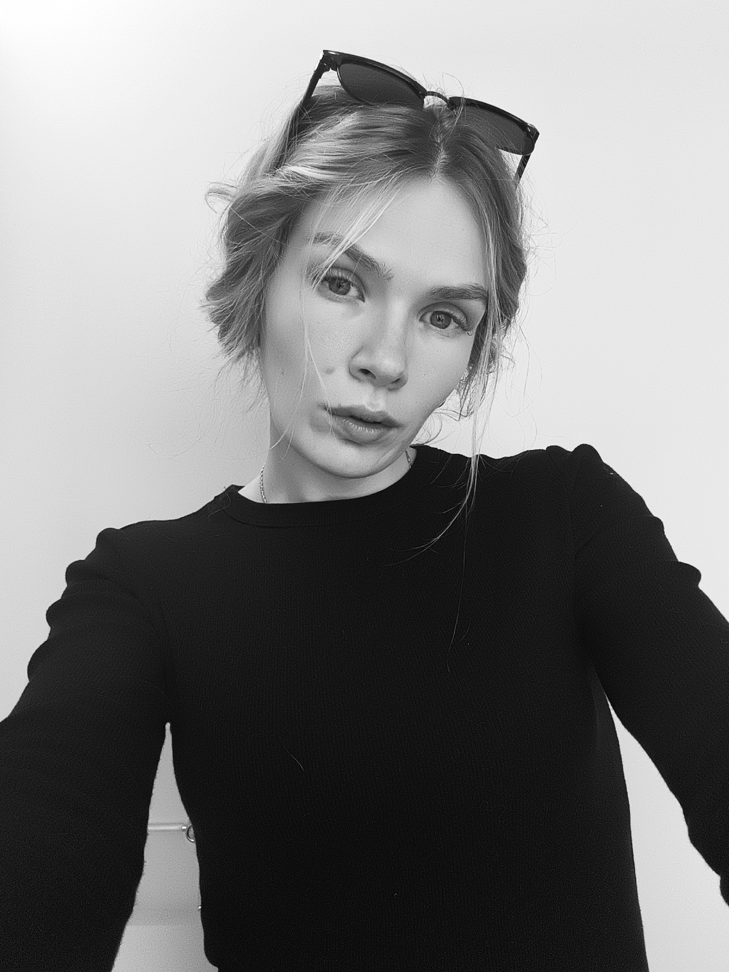 A black and white, low-angle selfie shows a young woman with light-colored hair, arranged in a loose updo, wearing sunglasses perched atop her head. She has light eyes and lips, and is wearing a dark, long-sleeved shirt. A thin chain necklace is visible. The background is a plain, light-colored wall. There is no information in the image to indicate the location is Izhevsk, Russia.
