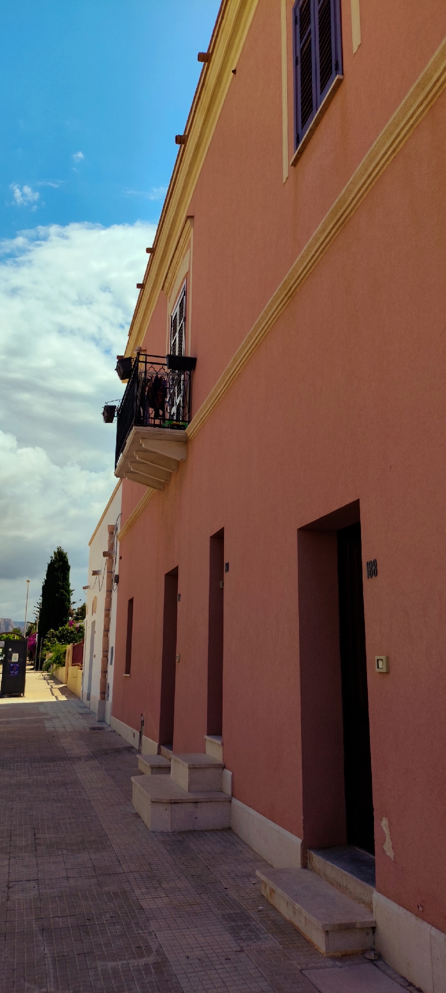 This is a vertical, eye-level, medium shot of a street in San Vito Lo Capo, Italy. The foreground is occupied by a tiled sidewalk. To the right, a coral-colored building with a light yellow trim features three recessed doorways. The number "138" is visible above the rightmost doorway. Steps lead up to each entrance. Above the building's ground floor, a balcony with black metal railings extends from the wall. The upper story has a window with dark shutters. In the background, a thin, dark tree stands next to a white building. Further back, flowering bushes and a distant mountain range are visible under a blue sky with scattered clouds. A black trash receptacle is present on the sidewalk to the left. The overall impression is one of a quiet, sun-drenched street.