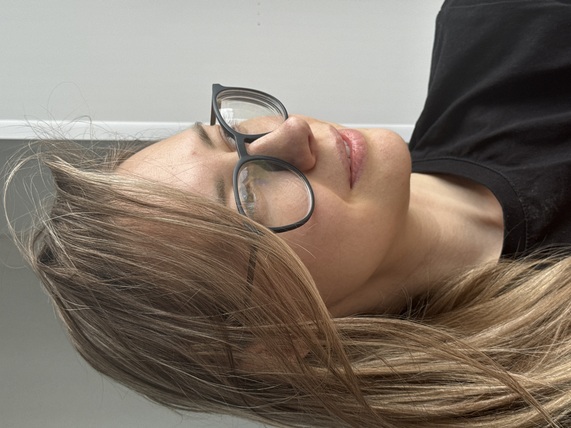 A close-up, tilted shot focuses on a fair-skinned woman with blonde hair and wearing glasses. She is lying down, with her head tilted back. The background is a light gray surface meeting a white wall. The woman is wearing a black shirt. The location is indeterminate.