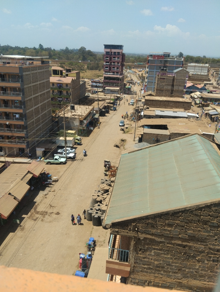 This elevated wide-angle image depicts a bustling street in the Witeithie ward, Kenya, under a clear sky with scattered clouds.
The central feature is a wide, unpaved dirt road, light brown in color, extending from the foreground into the distance. Utility poles with dense overhead wiring line both sides of the street.
On the immediate right, a building with a large, light green corrugated metal roof and exposed blockwork walls dominates the foreground, partially obscuring the street. Adjacent to it on the street level, a substantial pile of black cylindrical water storage tanks is stacked. Further down the street on the right, various commercial buildings with different finishes, including one multi-story structure with a grey-blue facade, are visible.
On the left side of the street, a multi-story building with a partially finished concrete facade and brown-railed balconies is prominent. Further along this side, several single and multi-story structures, some with brown awnings indicating market stalls, line the road. In the mid-distance on the left, a tall, slender multi-story building with a pink/maroon upper section and white-framed windows stands out.
Vehicles observed on or near the street include: a light green pickup truck and two white sedans parked on the left; a blue sedan and other vehicles parked or moving on the right; and at least one motorcycle.
Pedestrians are visible on the street. In the foreground, three individuals (two adults, one child) walk along the right side of the road. Multiple other small groups of people are scattered further down the street.
The background reveals a transition from urban development to a more natural landscape, characterized by a line of green trees and distant fields under a hazy horizon.