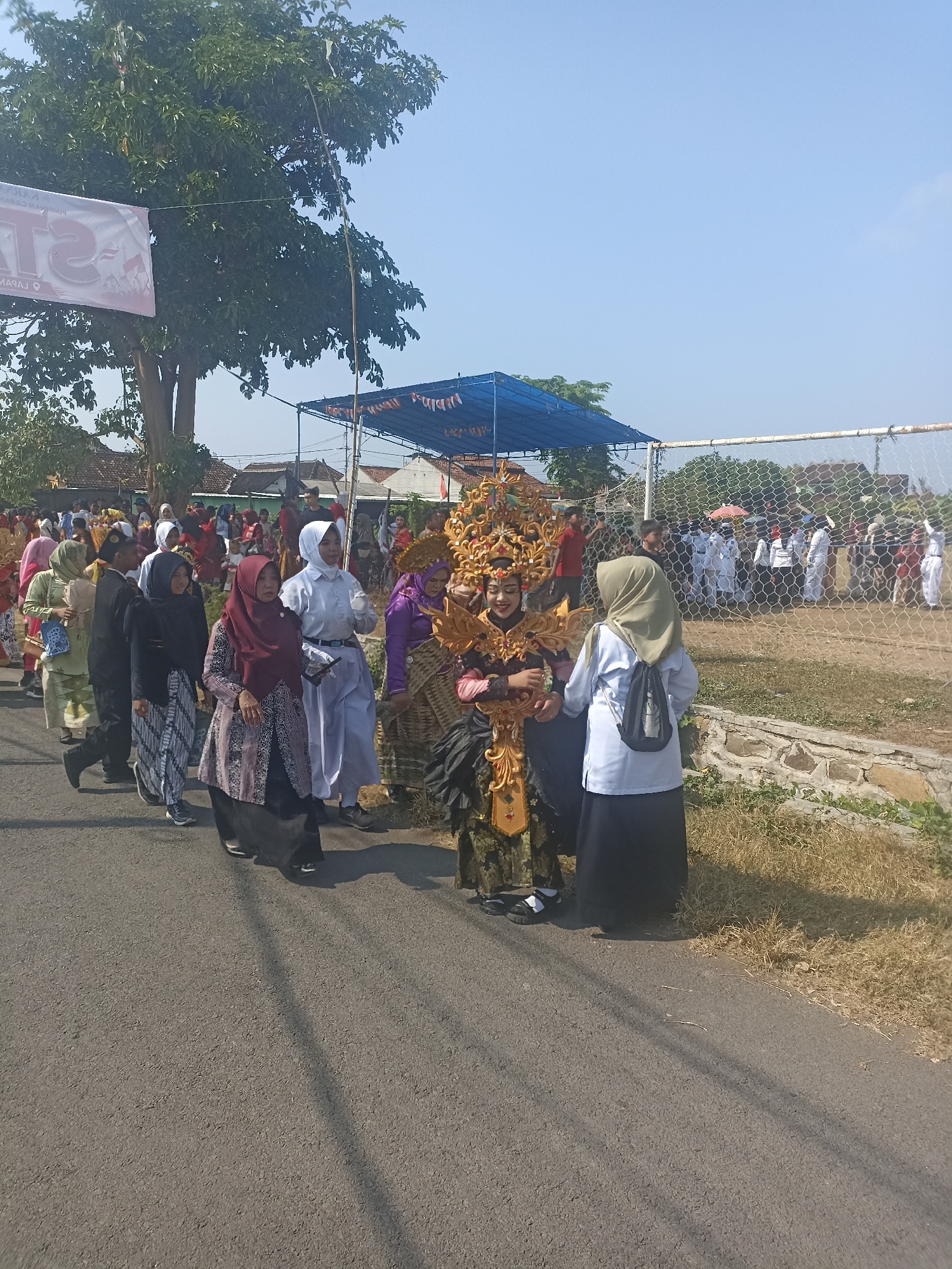 In Banyuwangi, Indonesia, a procession is underway along a paved road, with a group of people participating in what appears to be a cultural event or parade. A person in the foreground is adorned in an elaborate golden costume, featuring a headdress and intricate detailing on their garment. They are surrounded by other individuals, many of whom are wearing traditional Islamic attire, such as hijabs and modest clothing. The procession is moving from left to right in the frame, with spectators and participants lining the road.
In the background, a blue tent structure is visible, suggesting an organized event or viewing area. Beyond that, a white fence and a grassy field, possibly a sports ground, can be seen. The sky is clear and blue, indicating a sunny day. Trees and some buildings are present on the left side of the image, contributing to the suburban or semi-rural setting within Banyuwangi. The overall atmosphere suggests a festive and communal gathering.