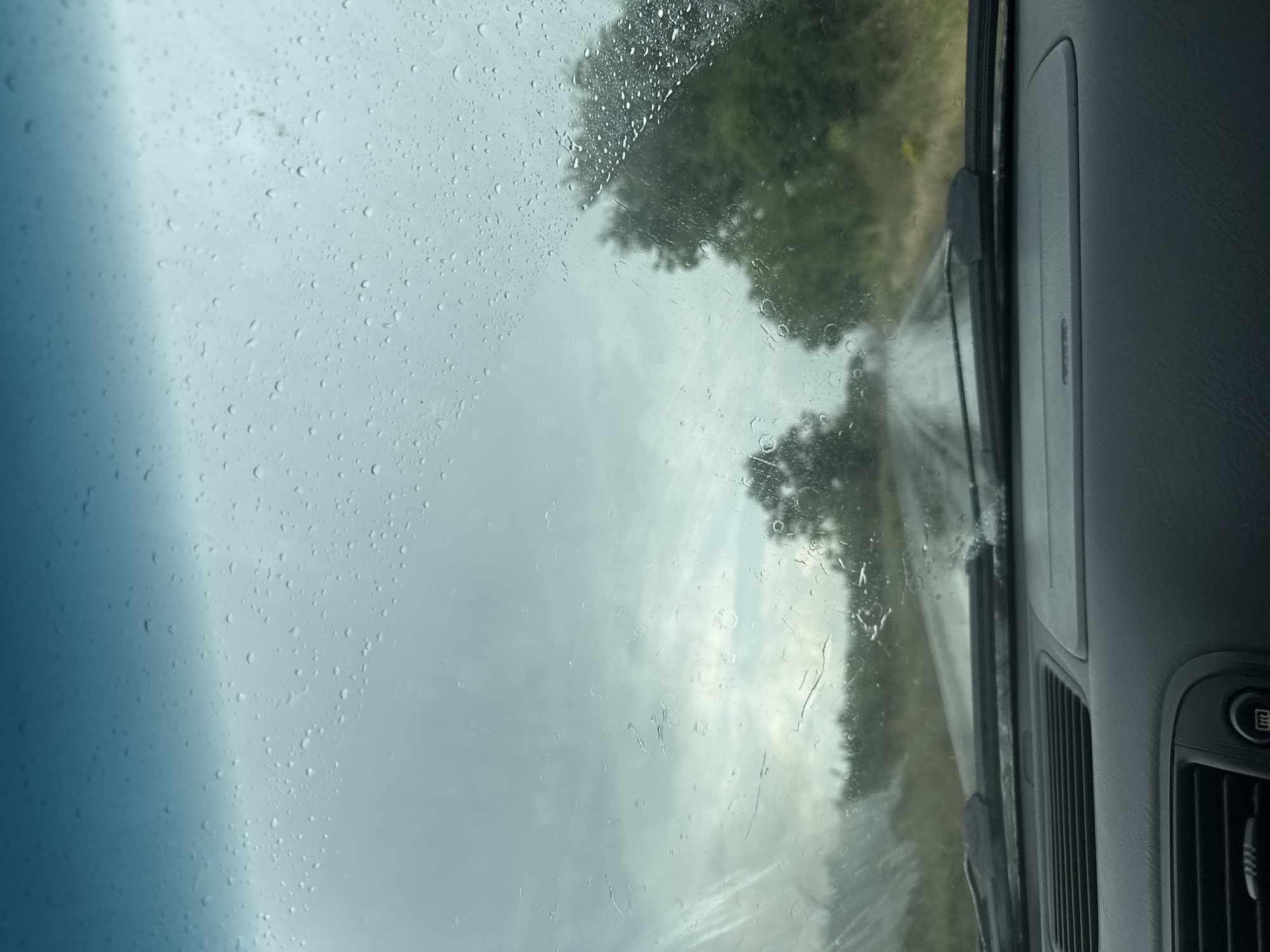 This image captures a view through a car windshield on a rainy day, looking out at a road leading through a rural landscape. The windshield is covered with numerous raindrops, some small and distinct, others larger and streaked, obscuring the view and distorting the scene beyond.
The road ahead appears wet and glistening, indicating recent or ongoing rain. The surrounding environment is characterized by lush green trees and vegetation on either side of the road. The sky is overcast and gray, typical of rainy weather, with some lighter patches suggesting diffused sunlight attempting to break through the clouds.
The dashboard of the car is visible in the foreground, showing parts of the car's interior. This includes air vents and the edge of the dashboard console. The windshield wipers are also visible, resting at the bottom of the windshield.
The image conveys a sense of driving in challenging weather conditions, where visibility is reduced due to the rain on the windshield and the overcast sky. The overall mood is somewhat somber and subdued, reflecting the weather. The location is specified as Cherni Vrah, Bulgaria, suggesting a natural, possibly mountainous or hilly, terrain commonly found in such regions.