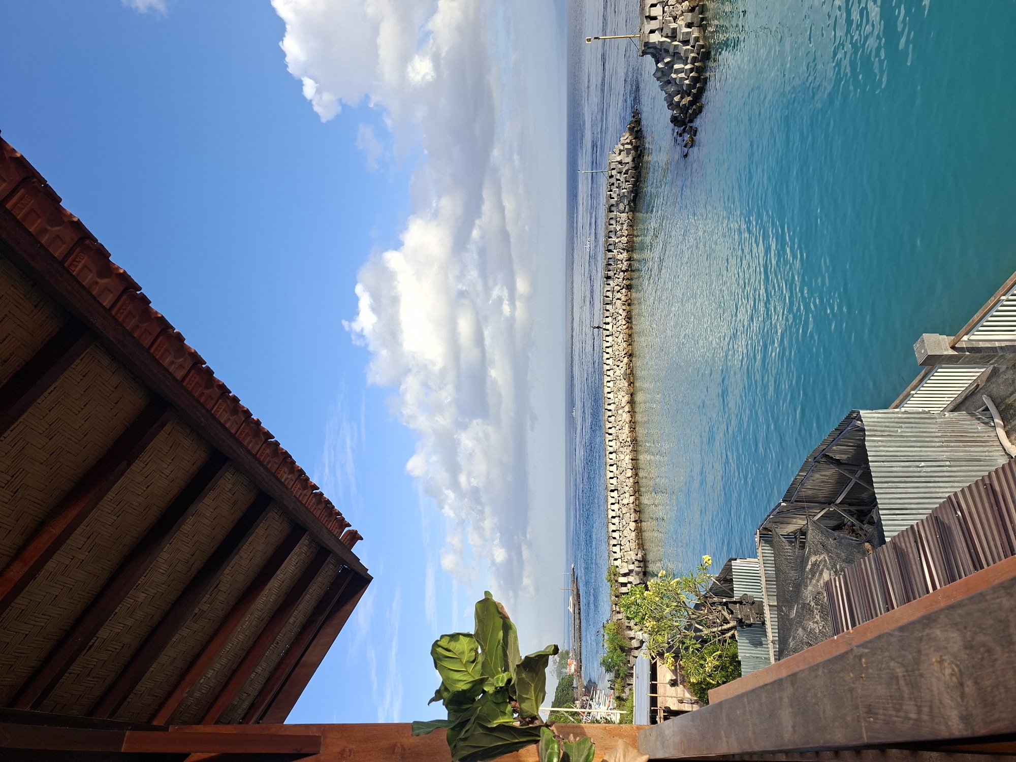 The image depicts a view from a wooden balcony overlooking a tranquil ocean harbor in Nusa Penida, Indonesia. The foreground is dominated by the wooden structure of the balcony, with a large green leafy plant on the left. The balcony overlooks a clear turquoise ocean, protected by a stone breakwater. Beyond the breakwater, the open sea stretches to the horizon under a bright blue sky dotted with white clouds. A few boats are visible on the water near the shore in the distance. The scene suggests a peaceful, seaside location, likely a resort or residential area, enjoying the morning light.