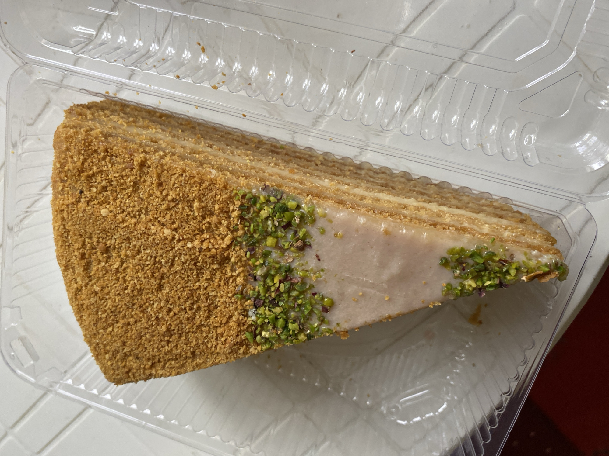 A slice of honey cake, topped with crushed nuts and pistachios, sits in a clear plastic container. The cake has multiple thin layers and a light-colored frosting. The container is on a white, textured surface, possibly a table. The scene is indoors.