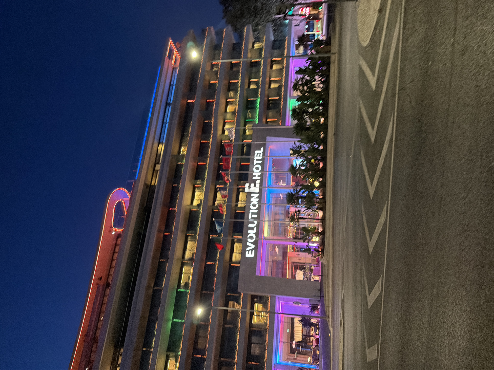 The image shows the "Evolution Hotel" in Cascais, Portugal, at dusk or night. The hotel is a multi-story building with modern architecture, illuminated by various colored lights. The main entrance is prominent, with large glass windows and colorful lighting in shades of pink, purple, and blue. Above the entrance, the hotel's name, "EVOLUTION HOTEL", is displayed in white illuminated letters.
Several flags are visible on flagpoles in front of the hotel. The building's facade features multiple floors with rows of windows, and some floors are accented with green and orange lighting. The rooftop is outlined with red and blue neon lighting.
In the foreground, a street with traffic markings is visible, suggesting the hotel is situated along a road or avenue. Palm trees and other plants are planted in front of the hotel, adding to the environment. The sky is dark blue, indicating it is nighttime or late evening. There are no discernible people in the image. The overall impression is that of a vibrant and modern hotel illuminated for the night.