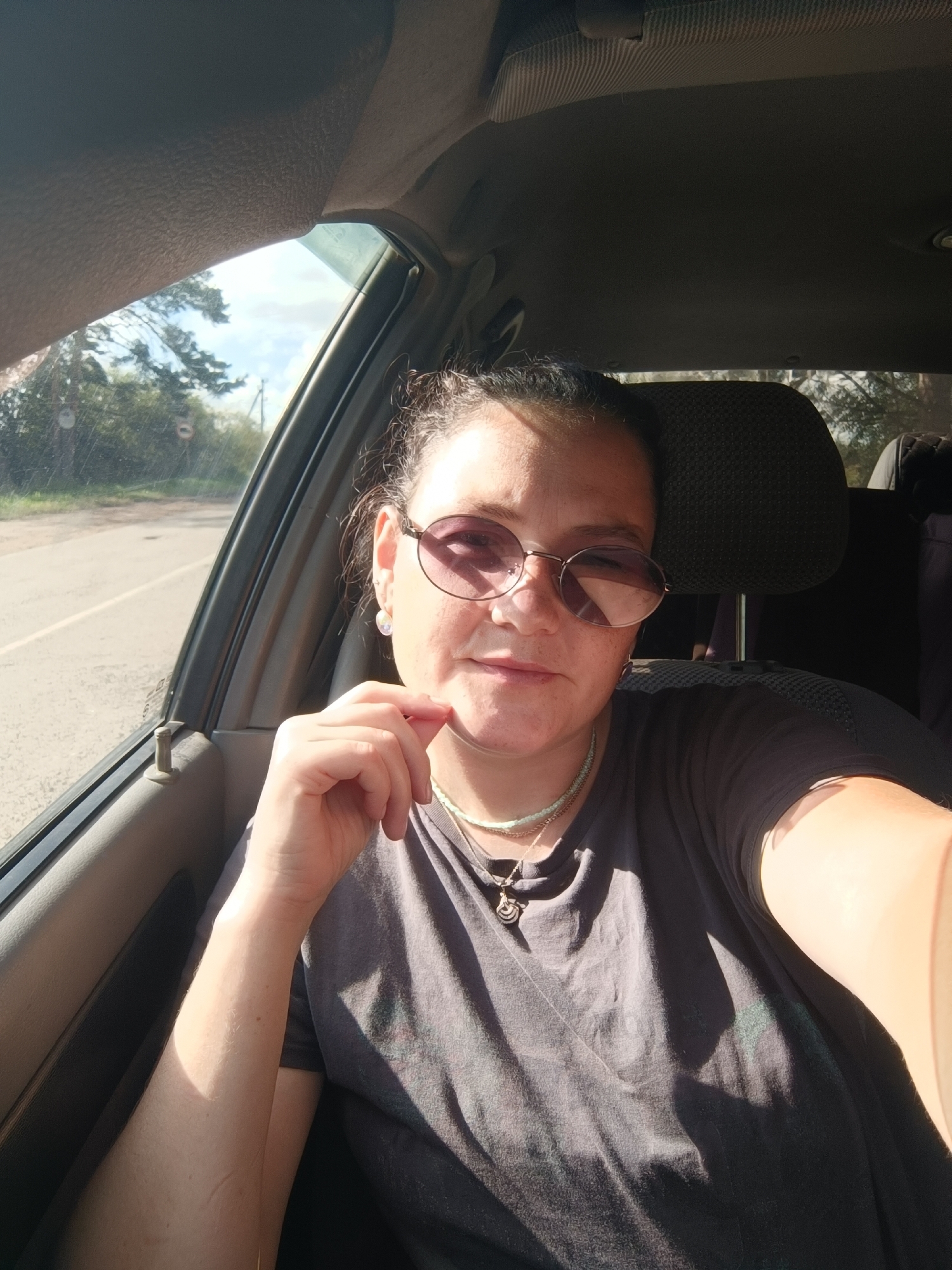 A medium-shot, selfie-style photograph taken from inside a vehicle, shows a woman with dark hair pulled back, wearing round, tinted sunglasses, and a dark gray t-shirt. She has her right hand raised to her chin, with her index finger touching her cheek. A necklace with a pendant is visible around her neck. The interior of the car, including a headrest and part of the dashboard, is visible in the background. Sunlight streams through the open window on the left, illuminating the woman and casting shadows. Outside the window, a road, trees, and sky are visible. The scene appears to be in a rural or semi-rural area, possibly on a road outside of Foki, Russia, given the presence of deciduous trees and a dirt or gravel shoulder along the road.