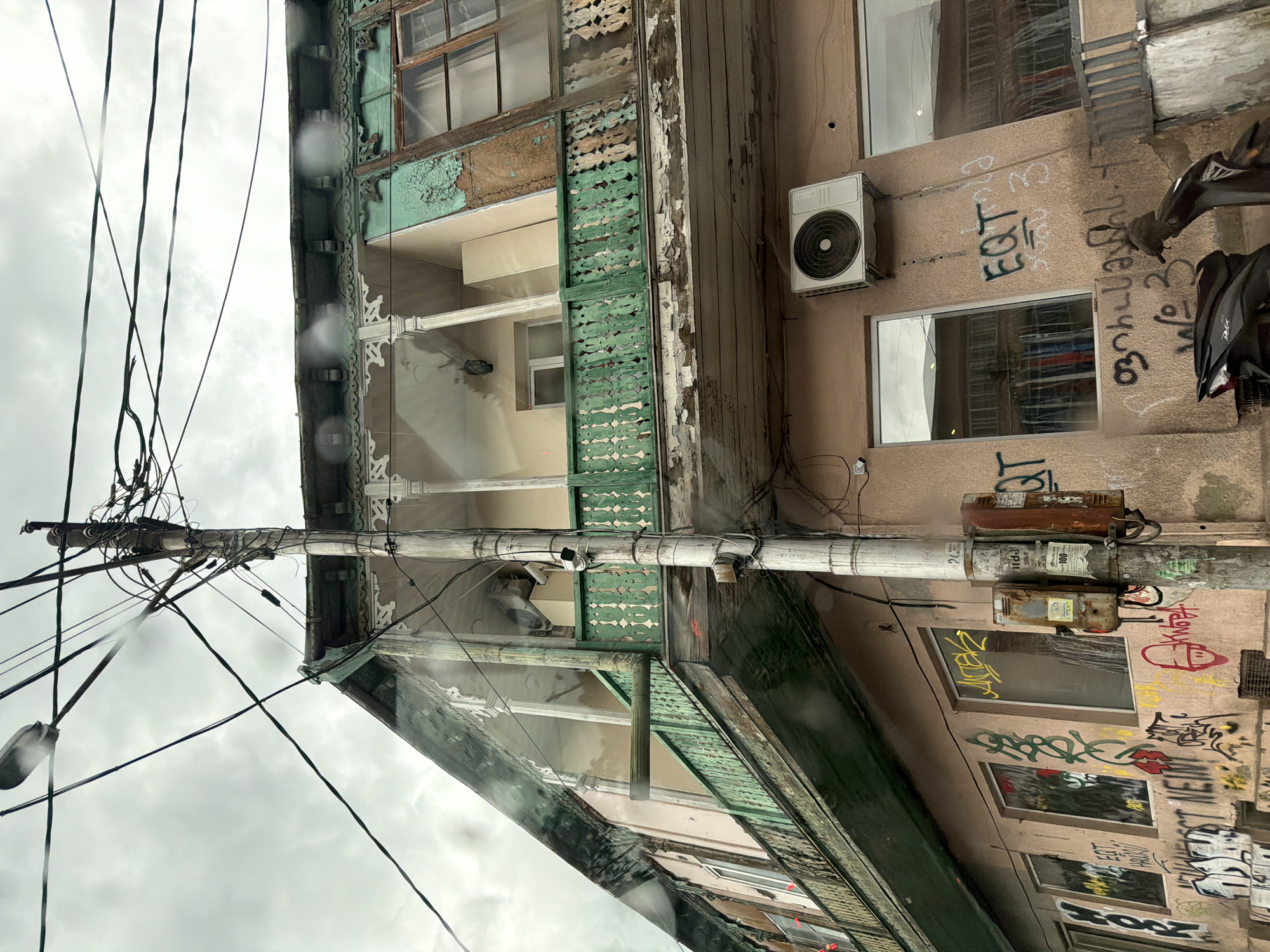 The image depicts an old, dilapidated building in Tbilisi, Georgia. The structure has peeling paint, especially in green and brown tones, and faded wooden details indicative of age and weathering. Balconies with decorative railings are visible. A prominent utility pole, tangled with wires, stands in front of the building. The exterior walls are covered with graffiti. An air conditioning unit is attached to one section of the building. A motorcycle is partially visible on the right side of the frame. The sky is overcast, suggesting rainy or cloudy weather.