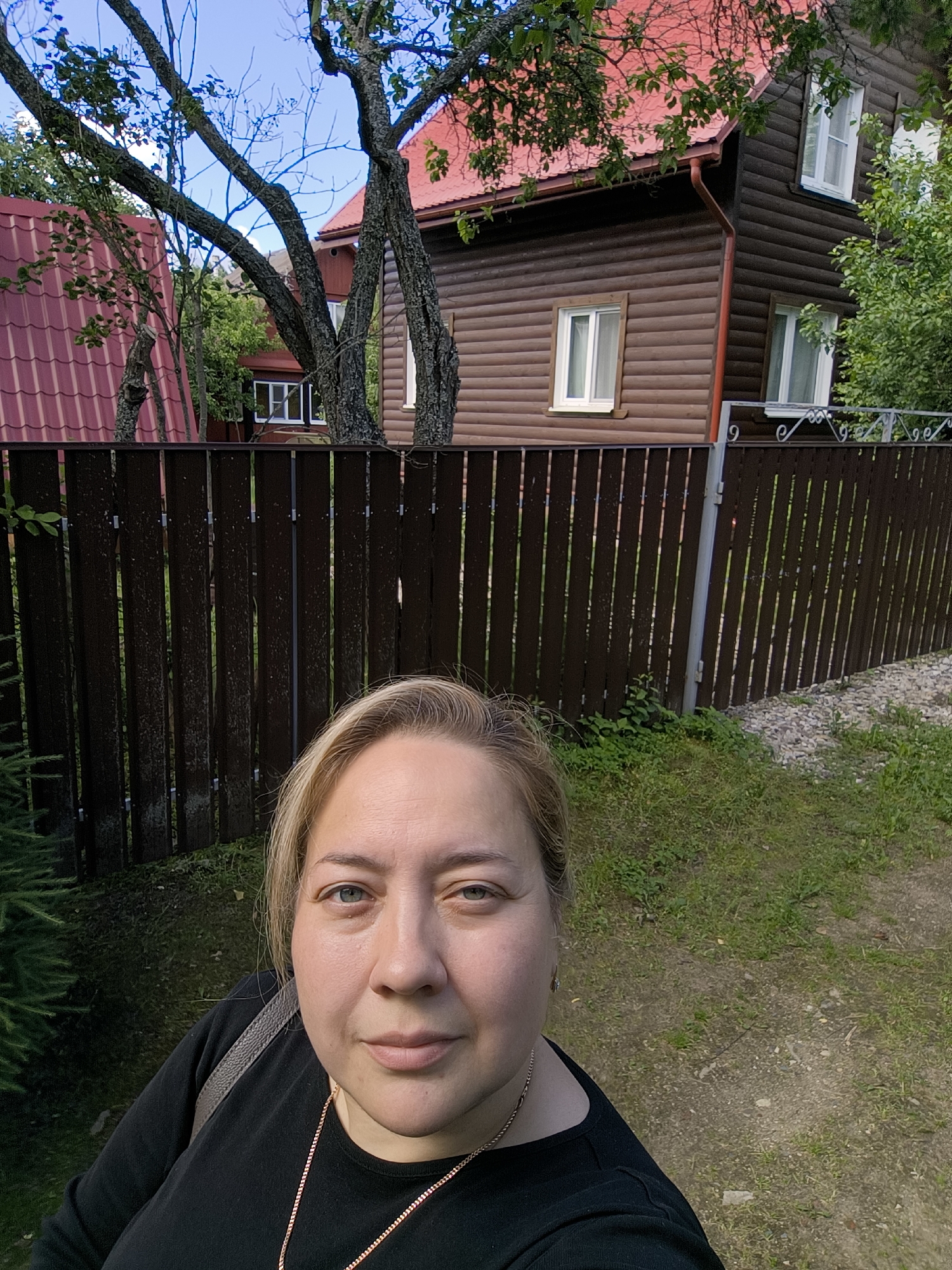 This is a selfie taken outdoors in Novonikolskoye, Russia. A Caucasian woman with blonde hair and blue eyes is in the foreground, wearing a black shirt and a gold necklace. She is looking at the camera. Behind her, a brown wooden fence separates the foreground from a residential area. Two wooden houses with red roofs are visible beyond the fence. The house on the right has a dark brown exterior with white-trimmed windows. The house on the left has a lighter brown exterior with a red metal roof. Trees with green leaves are scattered throughout the scene. The ground in the foreground is covered with grass and dirt, with some gravel on the right side. The sky is blue with some scattered clouds.