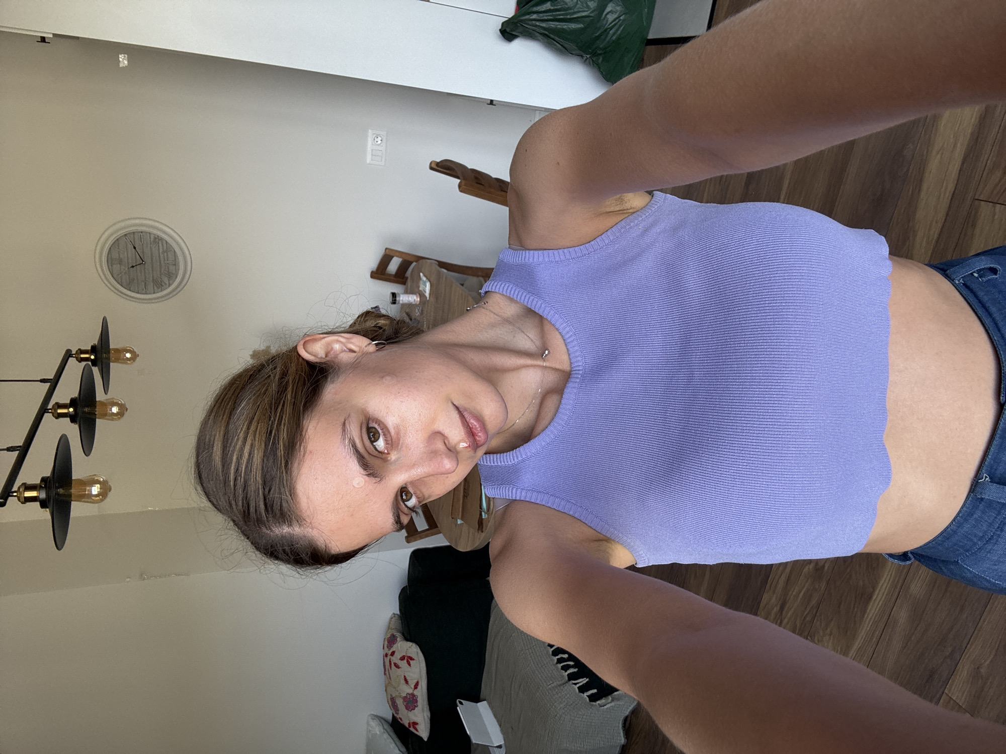 This image is a selfie taken by a woman indoors, capturing her face and upper body from an upward angle. The woman has brown hair, styled with some lighter highlights, and is looking directly at the camera with a neutral to slightly pensive expression. Small, clear droplets are visible on her forehead and upper lip. She is wearing a light purple, ribbed sleeveless crop top and blue denim jeans, along with a delicate silver necklace.
The background suggests a home interior, possibly a living or dining area. A light-colored wall features a decorative circular clock with a gray, wood-textured face and black hands. To the left, a black chandelier-style light fixture with visible Edison bulbs is partially shown. Wooden furniture, including chairs, can be seen in the background. The floor is made of dark wooden planks. Other elements include an electrical outlet on the wall, a dark green bag partially visible in the upper right, and a couch with patterned pillows and a throw blanket towards the bottom right. The lighting indicates it is daytime. No visible text is present in the image.