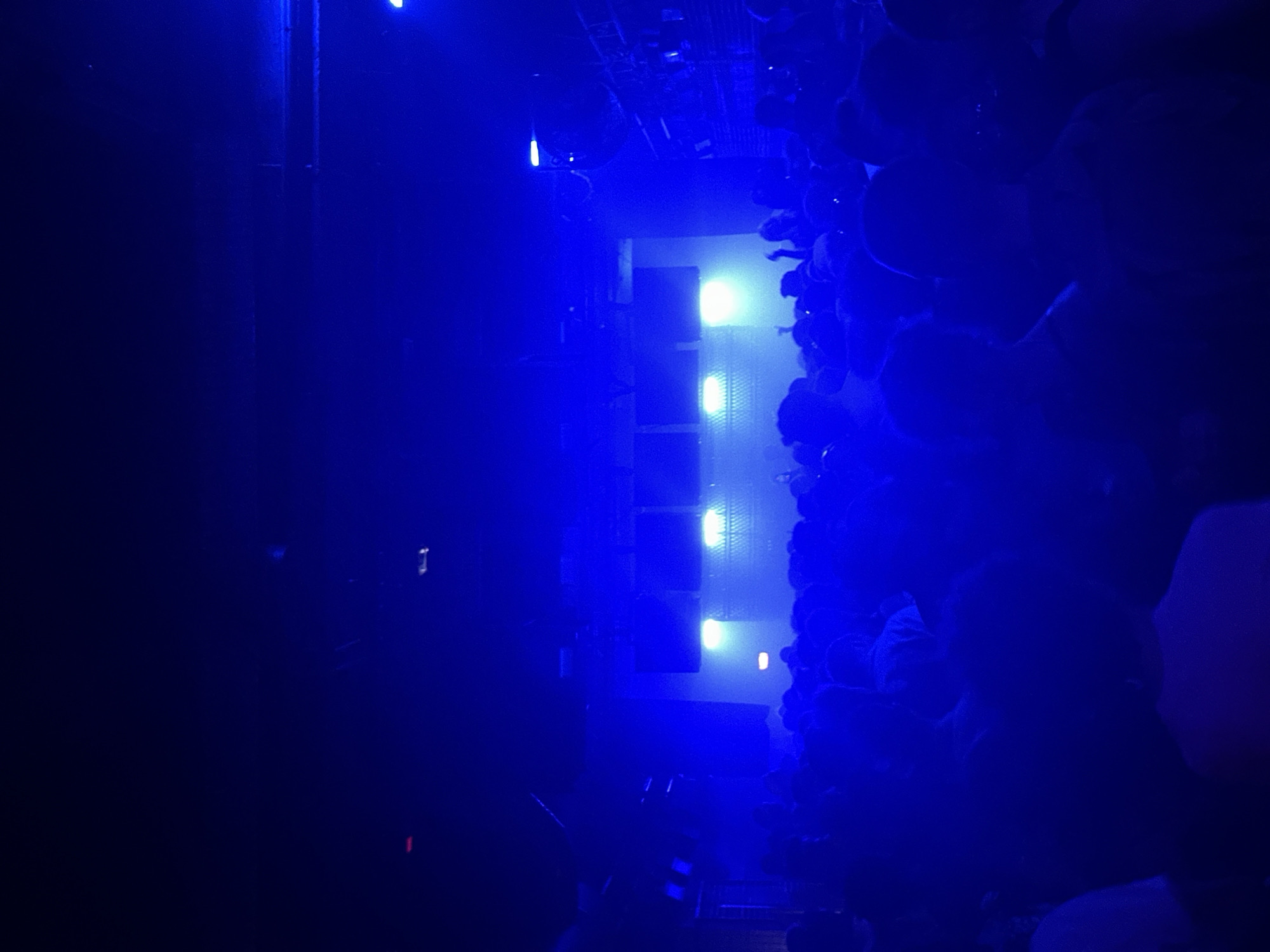This media file captures an indoor event, likely a concert or performance, in Paris, France, characterized by a dominant artificial blue lighting scheme. The image is taken from a slightly elevated perspective, looking down and across a darkened venue.
On the left side of the frame, a large section of the venue is mostly dark, with only faint glimmers or reflections suggesting distant elements. The central part of the image features a focal point or stage area, equipped with several bright, rectangular light fixtures arranged vertically, emitting a yellowish-white glow. Adjacent to these lights are large, dark structures that appear to be speaker stacks or other performance equipment.
To the right, a dense crowd of people is visible, mostly as dark silhouettes against the blue light. Their forms are clustered together, indicating an engaged audience at a live event. The pervasive blue light creates a cool, immersive, and energetic atmosphere, typical of a nighttime concert or club setting. The activity is clearly a gathering for a performance, likely a musical act or DJ set.
The time of day is certainly night, or an equivalent indoor setting where natural light is absent. There are no discernible weather conditions as it's indoors, and no visible text can be identified in the image. The overall mood is one of excitement and immersion in the performance.