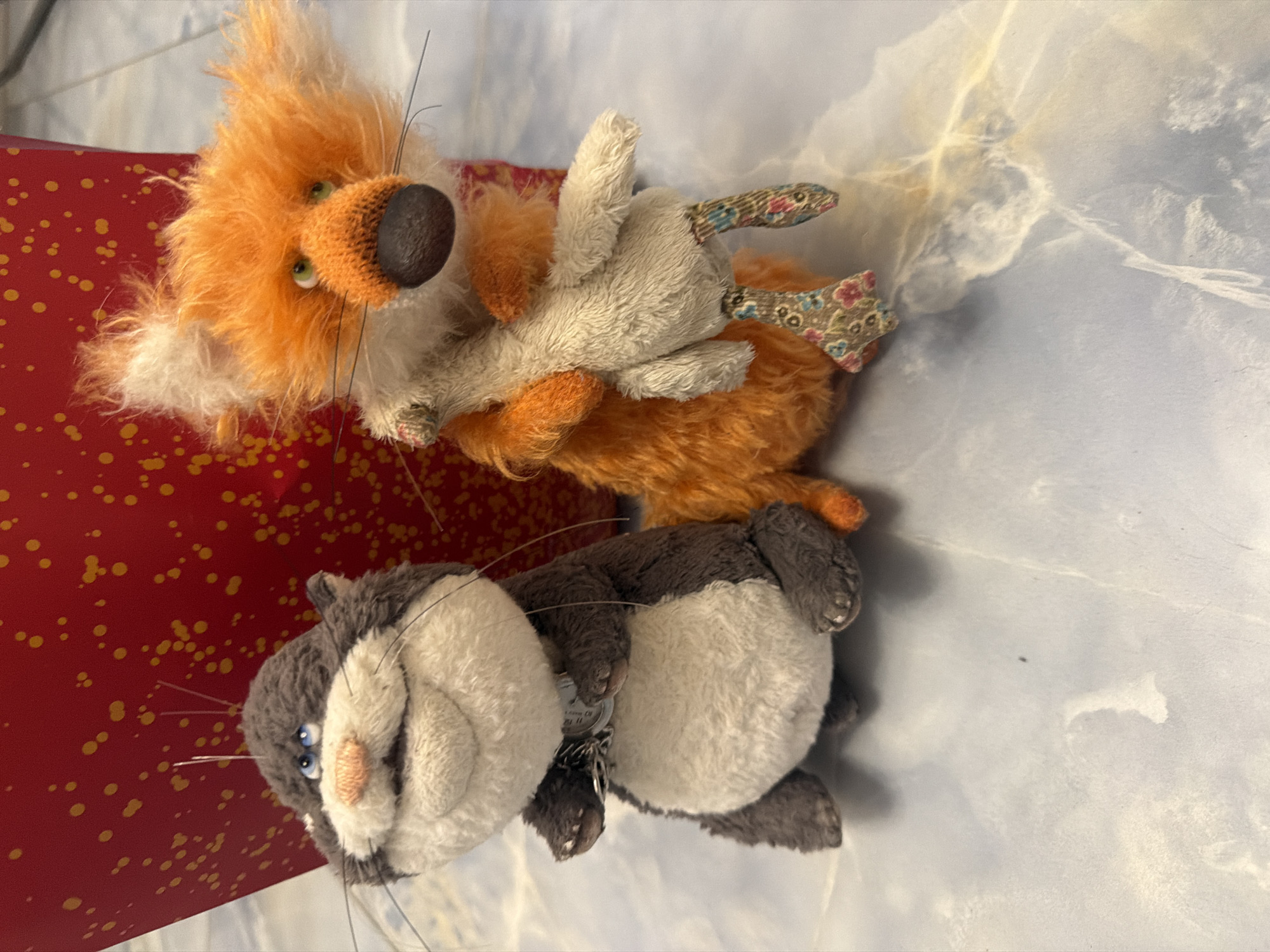 Two stuffed toys, a gray and white cat-like creature and an orange fox-like creature, are positioned on a light blue surface with a marble-like pattern. The cat toy is on the left, facing forward with its mouth open and eyes looking up and to the left. It has a light gray body with a white belly and a dark gray head and back. A silver chain or necklace is around its neck. The fox toy is on the right, also facing forward, with its head tilted slightly to the left. It is predominantly orange with white fur around its face and chest, and it is holding a small, light-colored stuffed animal. The background is a bright red object with yellow speckles. The scene appears to be indoors, on a flat surface, possibly a table or floor.