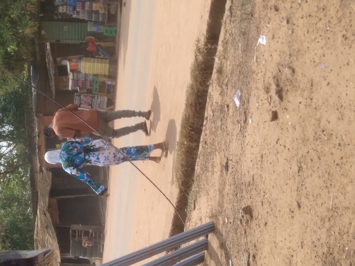 The image displays an outdoor street scene in Agege, Nigeria, under bright daylight. The foreground consists of exposed reddish-brown dirt, irregularly shaped small rocks, and scattered pieces of white debris. On the far left, a stack of grey, cylindrical pipes is positioned vertically. A thick black cable extends diagonally from the lower left quadrant towards the upper right, passing in front of two individuals.
Beyond a narrow, shallow dirt trench or culvert, two people walk away from the camera on a light-colored, unpaved road or path. The individual on the left, appearing to be female, wears a white head covering (hijab) and a full-length, long-sleeved dress with a prominent blue, white, and possibly green abstract or floral pattern. She wears light-colored flat sandals. The individual on the right, appearing male, wears an orange-brown t-shirt, grey trousers, and dark sandals or flip-flops. He holds a small, dark object in his right hand, possibly a mobile phone.
In the background, several single-story commercial structures with corrugated metal roofs line the road. The structures appear aged or in disrepair. On the left side of the background, a shop entrance shows stacked tires. On the right, another shop features a black and white "POS" (Point of Sale) sign and multiple crates containing bottled beverages (soft drinks and water) stacked outside. A person wearing a red shirt is seated on a chair further back, near the shop displaying beverages. Green foliage and trees are visible in the distant background. The overall environment appears dusty.