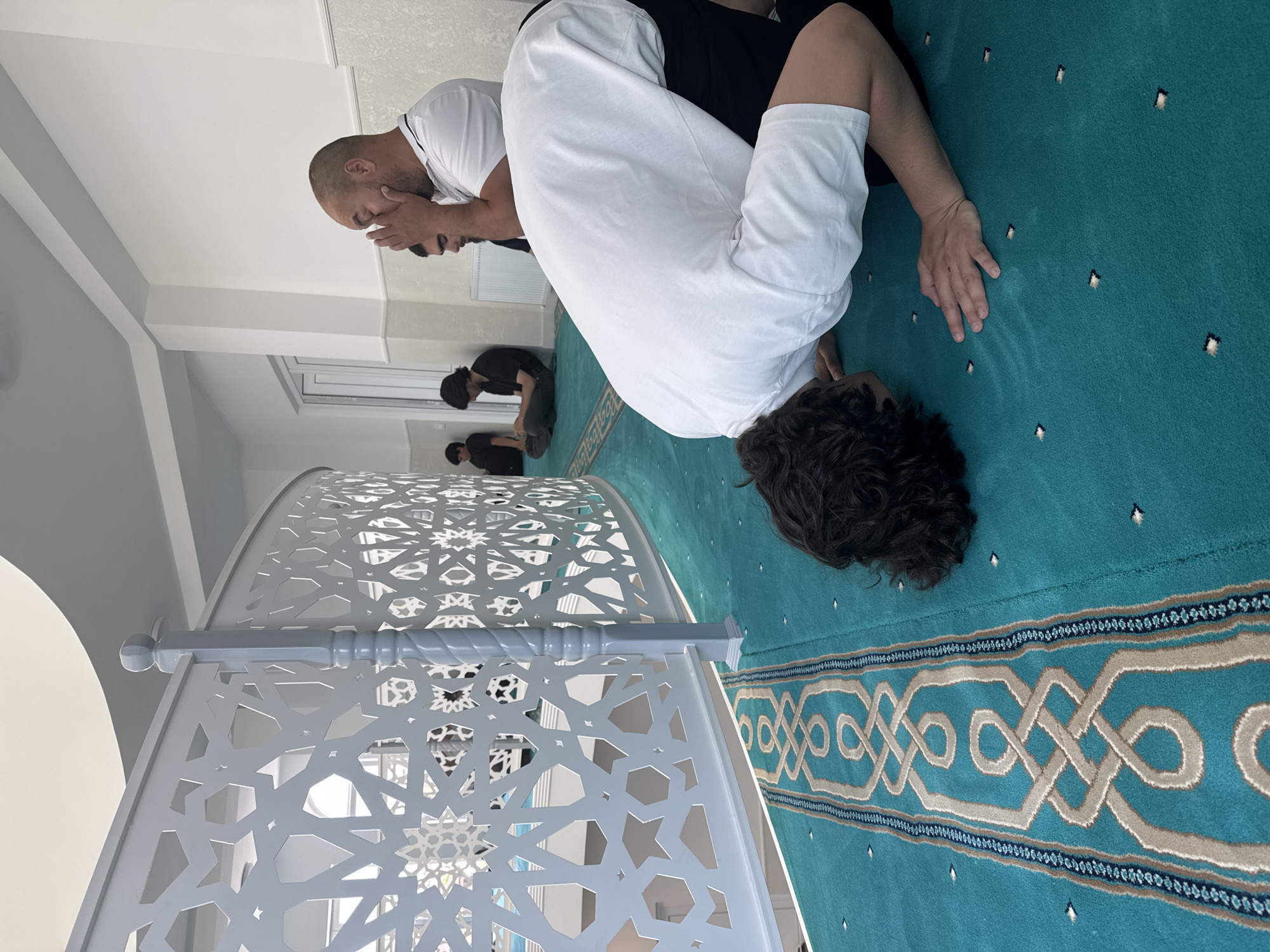 The image depicts several people engaged in prayer on a teal carpet in what appears to be a mosque. The camera is oriented vertically. In the foreground, a person wearing a white robe is in a prostrated position, with their head bowed. To their left, another person in a white t-shirt is also praying, their hands covering their face. Further in the background, two more individuals are visible in similar prayer postures. The setting features a white, intricately patterned decorative screen on the left, and a light-colored wall and ceiling. The teal carpet has a repeating geometric pattern border. The scene suggests a moment of religious devotion. The lighting appears to be artificial, possibly from overhead lights, as the environment is brightly lit without visible natural light sources like windows or sunlight. There is no visible text in the image. The location context of Novaya Adygeya, Russia, indicates the geographical setting of this religious gathering.