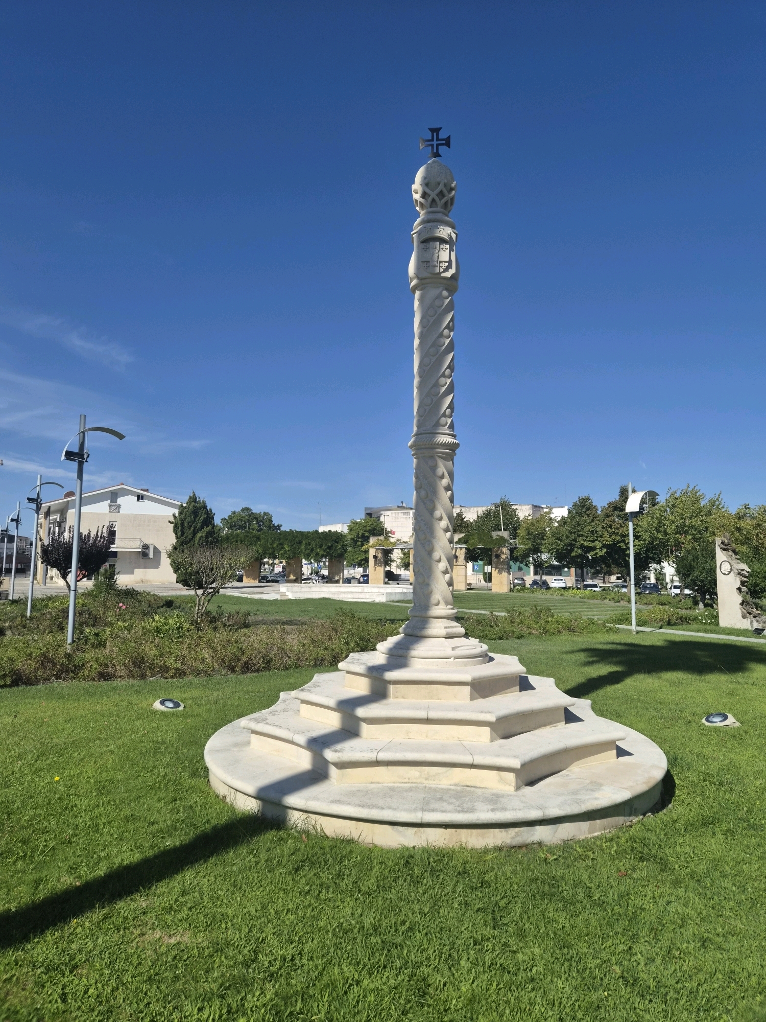 This is a photograph of a stone monument topped with a cross, located in a park in Batalha, Portugal. The monument is white stone with intricate carvings, including a spiraling pattern on the shaft and what appears to be a coat of arms or emblem near the top. The monument sits on a tiered, stepped stone base, which is surrounded by a well-maintained green lawn. In the background, there are trees, buildings, streetlights, and parked cars, suggesting an urban or suburban setting. The sky is a clear, bright blue, indicating a sunny day. The overall scene is peaceful and static, with no people or animals visible. The focus is on the monument itself, which is the central subject of the image.
