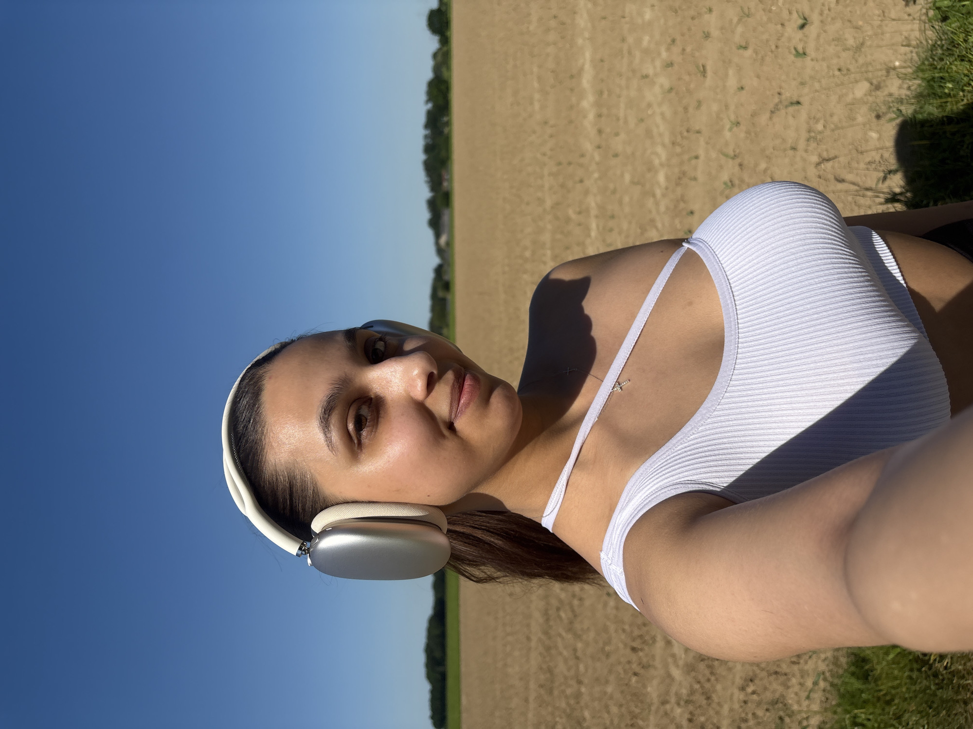 A young woman poses outdoors. She has brown hair pulled back with a white headband and wears white wireless headphones. She's wearing a white ribbed tank top with thin straps and a gold cross necklace. The background shows a clear blue sky, a tilled field, and a line of trees in the distance. The location could be an agricultural area near Viersen, Germany, given the landscape characteristics.