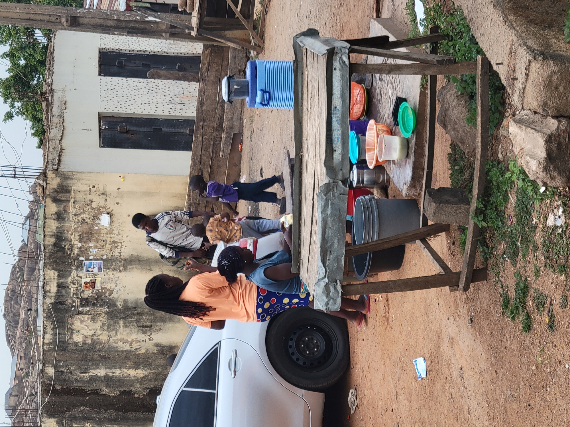 This outdoor scene in Jos, Nigeria, shows a makeshift water stand with several buckets and a cooler. Two women and a child are gathered at the stand, and another child is walking by. A man is sitting against a weathered wall in the background. The wall has posters on it. A portion of a silver car is visible in the foreground. The ground is dirt, and there are some rocks and vegetation present.