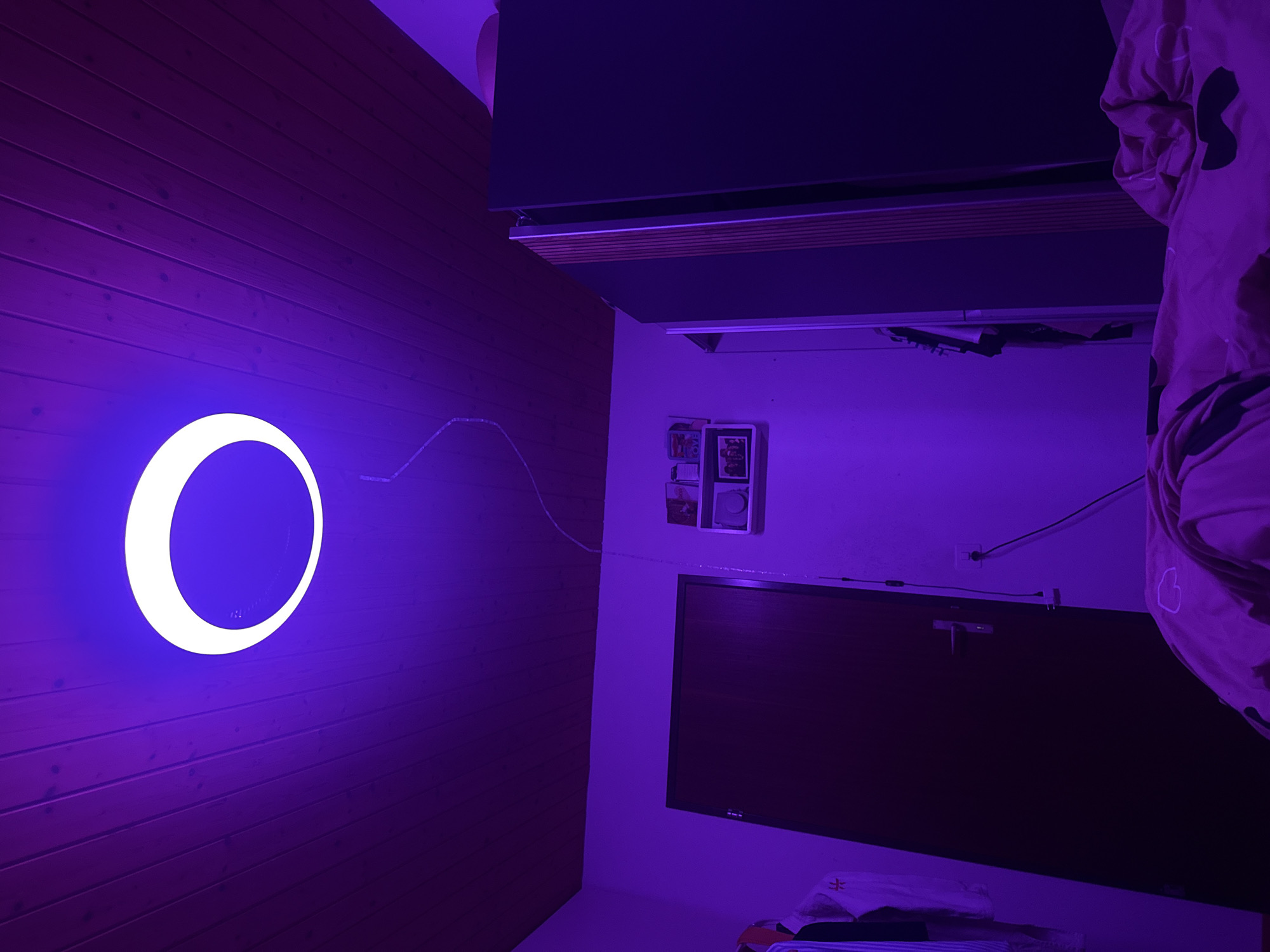 This image depicts an indoor bedroom or living space located in Oberflachs, Switzerland, illuminated primarily by a striking purple light. The ceiling is constructed from dark reddish-brown wooden panels, oriented diagonally across the frame from top-left to top-right.
Dominating the upper center of the image is a circular LED light fixture suspended from the ceiling. This fixture features a prominent outer ring of bright white light surrounding a central inner circle that emits an intense purple glow, bathing the entire room in a vibrant violet hue. A thin, translucent cable extends from this ceiling light, trailing vertically downwards along the wall to the right of the door.
The walls, appearing light-colored (likely white or off-white) under normal illumination, are saturated with the purple light. On the left side of the room, partially visible, are hanging clothes, including a white garment with a small red insignia. Centered on the back wall is a dark brown wooden door with a silver horizontal handle, which is closed.
To the right of the door, a small, two-tiered white wall shelf is mounted. It holds several indistinct objects, including what appear to be framed photographs and small decorative items. Below the shelf, a white electrical outlet is visible on the wall, with a dark cable plugged into it, extending downwards out of frame. The translucent cable from the ceiling light runs past this shelf and outlet.
On the right side of the room, a tall, dark-colored wardrobe or cabinet is partially visible. One of its doors is slightly ajar, revealing a narrow vertical strip of contrasting material, possibly light wood or a textured panel. In the upper right corner, behind the wardrobe, a soft, rounded pink or light purple object (possibly a cushion or a beanbag chair) is partially seen.
In the foreground, at the bottom of the image, rumpled bedsheets or a duvet in a pinkish-purple color with darker, irregular patterns (possibly hearts or abstract shapes) are visible, indicating the presence of a bed. No individuals are present in the scene.