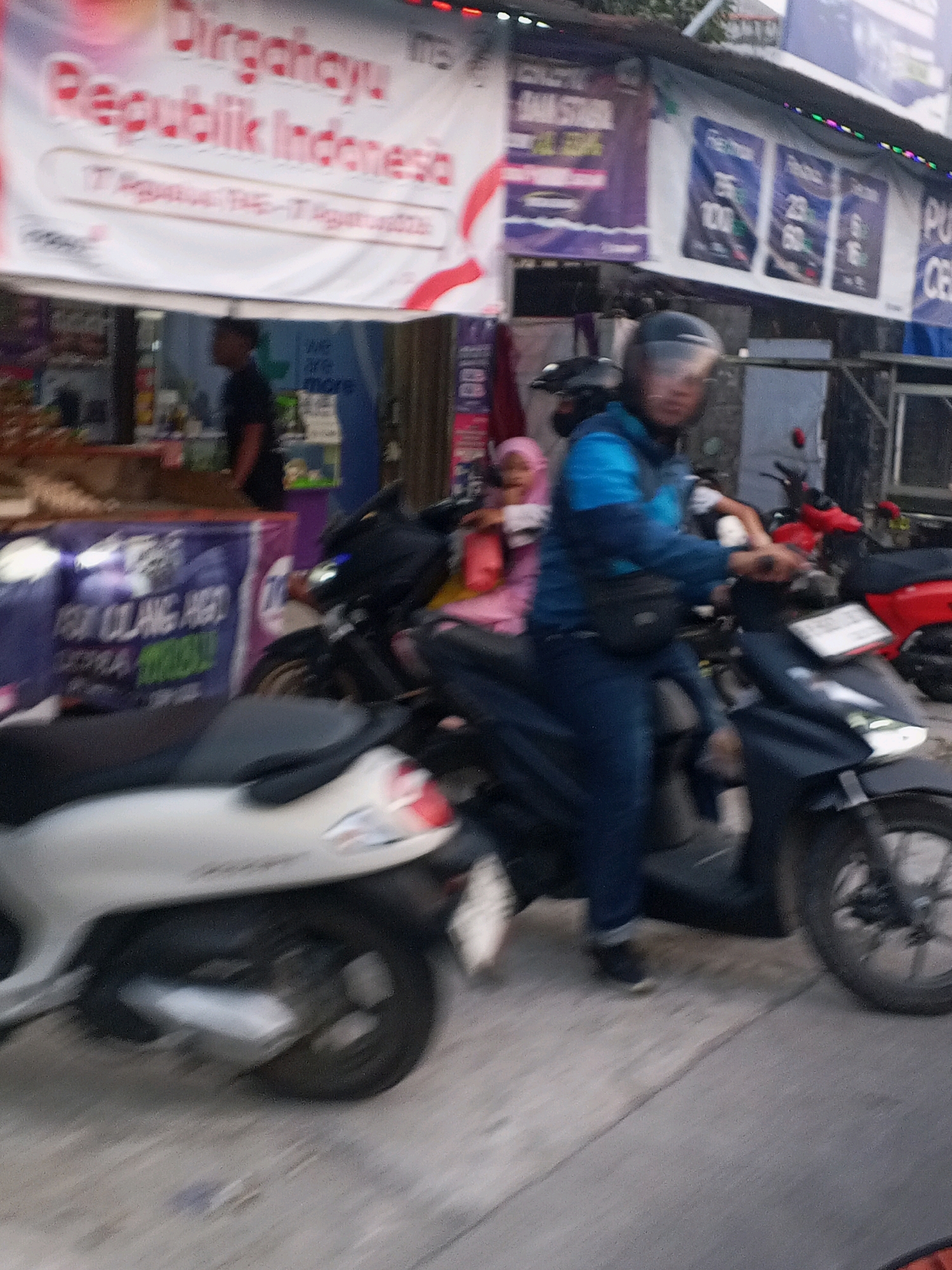 The image depicts a street scene in Ciruas, Indonesia, likely on or around August 17th, as indicated by a banner with the Indonesian text "Dirgahayu Republik Indonesia" and "17 Agustus 1945 - 17 Agustus 2023". A person wearing a blue jacket and a black helmet is riding a dark-colored motorcycle. Another person, a child wearing a pink hijab and pink clothing, is also visible on a motorcycle behind the first rider. In the background, there are several shops or stalls with various posters and banners displayed. One banner prominently features the red and white Indonesian flag design. Another banner in the background has purple and white text. Two other motorcycles are visible in the scene, one red and another white, with their rear ends partially visible. The ground appears to be paved. The overall impression is of a busy street with active motorbike traffic.