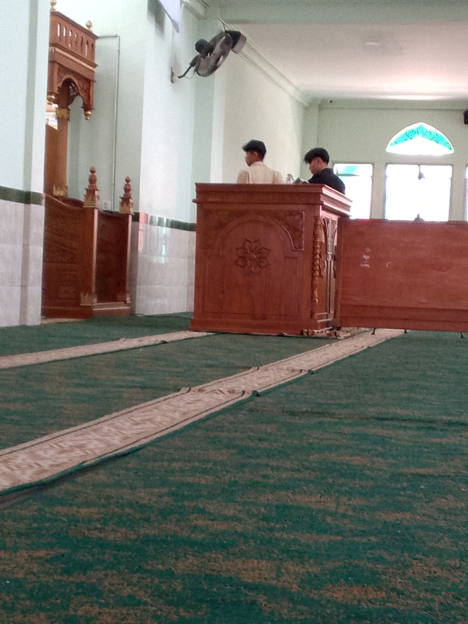 The image depicts the interior of a mosque in Cimahi, Indonesia, likely during prayer or a gathering. Two young men, one wearing a light-colored shirt and the other a dark shirt, stand behind a wooden pulpit. The pulpit is ornately carved with floral designs. In the background, a decorative wooden structure, possibly a minbar, is visible, along with white tiled walls and a fan mounted on the ceiling. Green carpet covers the floor, with a lighter-colored runner leading towards the pulpit. The overall scene suggests a peaceful and spiritual atmosphere within the mosque.