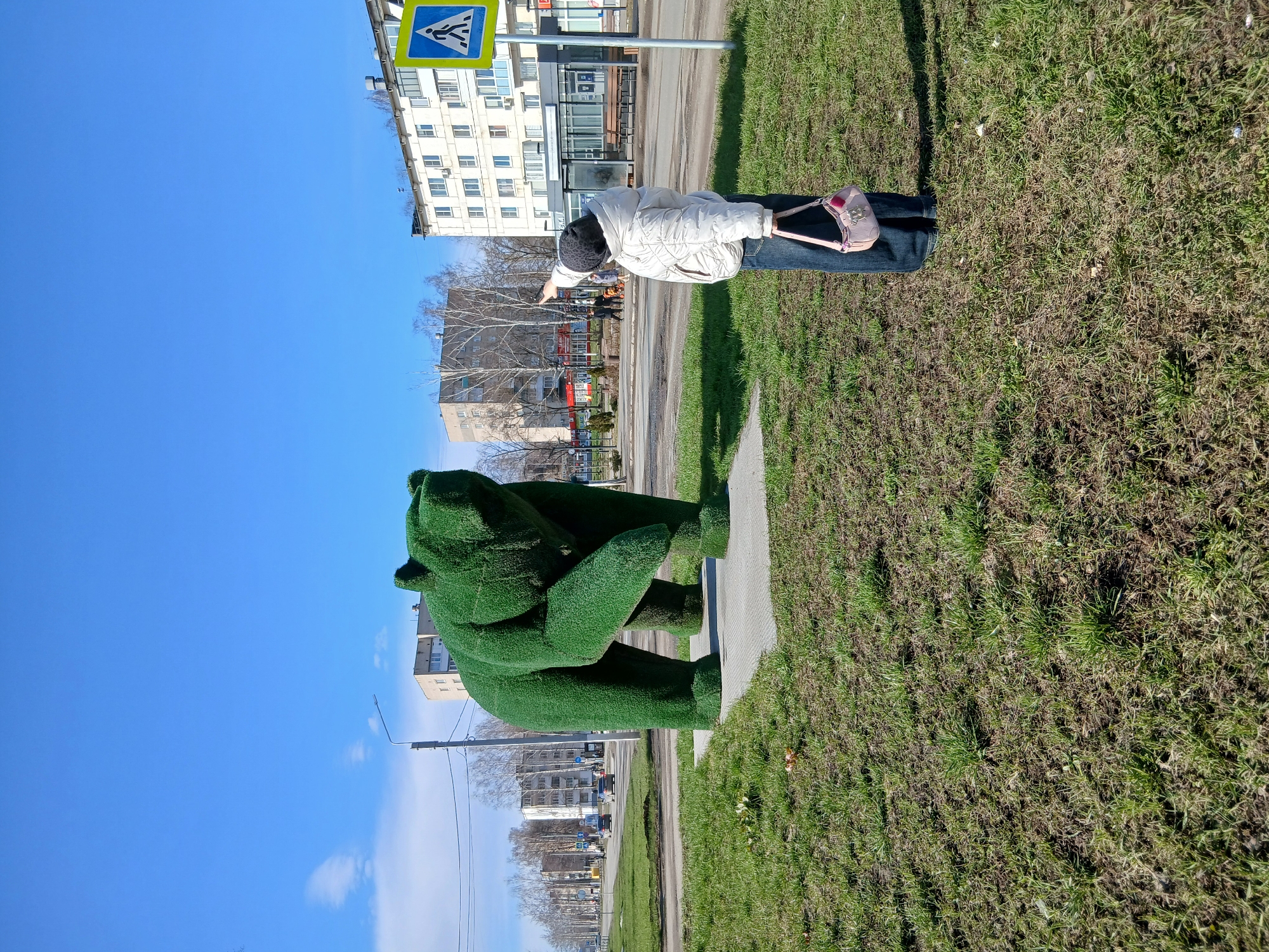 The image shows a person standing on a sidewalk next to a green grass sculpture of a bear, pointing toward a building. The location appears to be a city with a street running alongside the sidewalk and grassy area. A pedestrian crossing sign is visible near the person and building. The person is wearing a white jacket, blue jeans, and a dark hat, and carrying a pink purse.