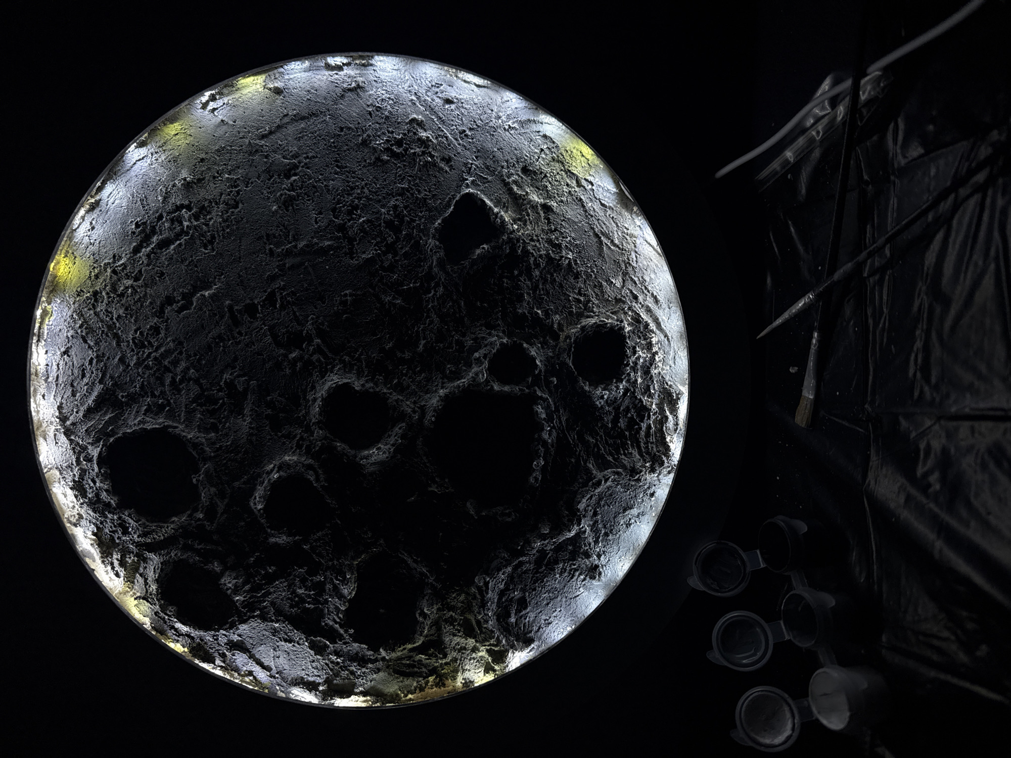 A close-up studio shot in Chaikovsky, Russia, depicts a model of a celestial body, possibly the moon or another cratered planet, with distinct dark and light regions, suspended against a black background. The model is illuminated, with a glowing edge and textured surface featuring numerous dark craters. In the right corner of the frame, partially obscured by shadow, are paint containers and a paintbrush, suggesting that the model is a creation of an artist and it is a work-in-progress. A dark, folded material or backdrop hangs vertically nearby.