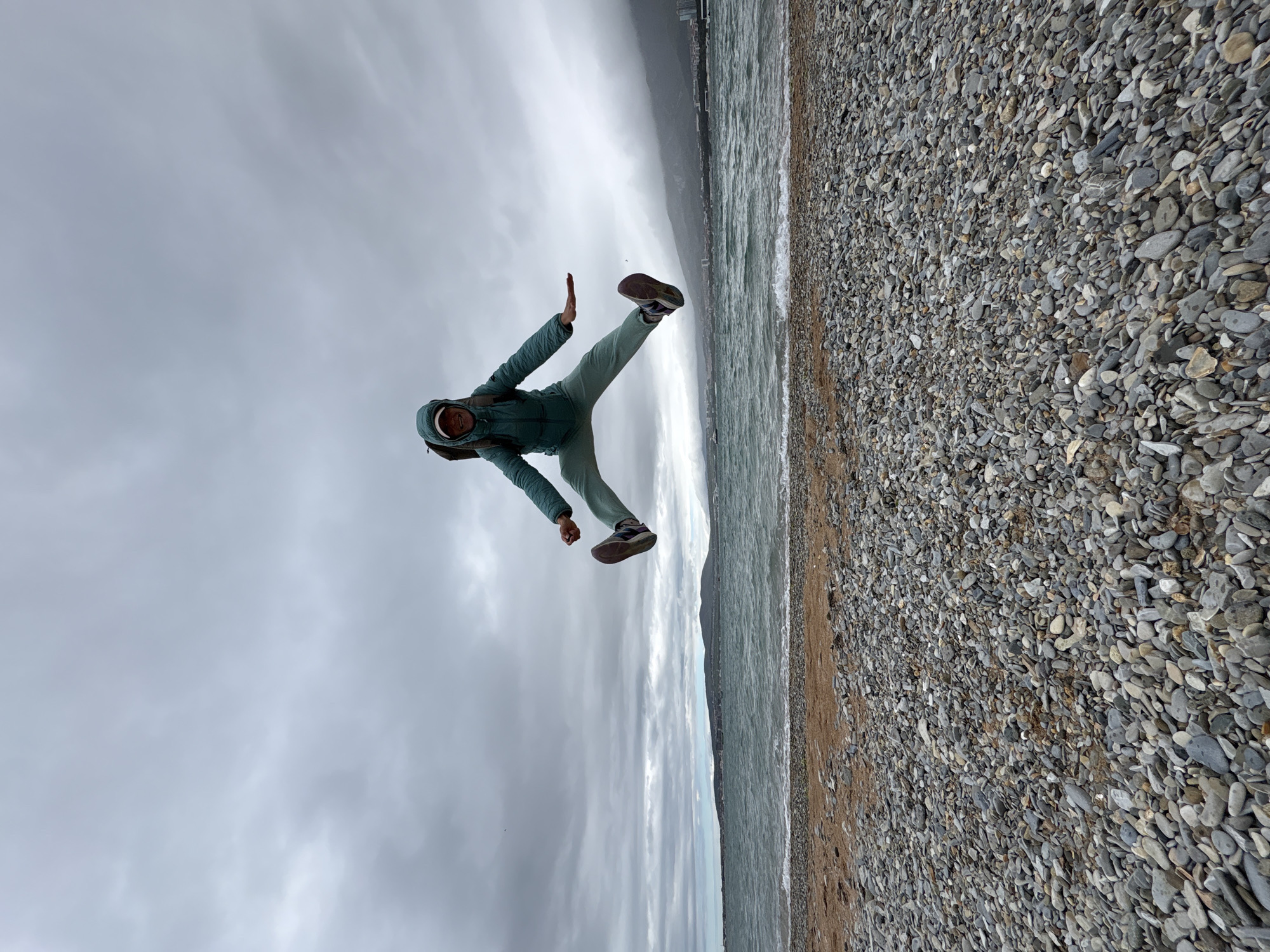 A person is jumping mid-air on a beach. The person is wearing a hooded jacket and pants, with shoes on their feet. They are positioned diagonally in the frame. The beach is covered in pebbles and small rocks, extending to the right side of the image. The ocean is visible in the background, with waves and a city skyline faintly discernible in the distance. The sky is overcast and cloudy. The location is Unknown, Unknown.