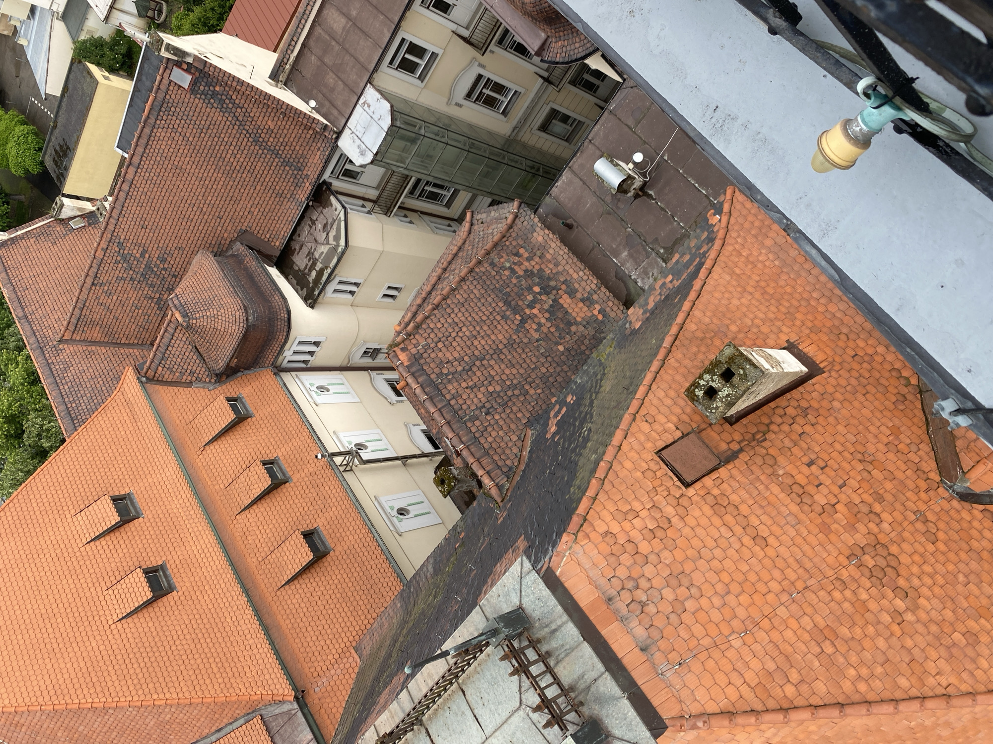 This is an aerial photograph of rooftops in the city of Bílina, Czechia. The scene is dominated by terracotta roof tiles, characteristic of European architecture. Several buildings are visible, including a prominent one with multiple dormer windows on its steeply pitched roof. Adjacent structures feature a mix of tile and darker, flat roofing materials. A modern glass-enclosed elevator shaft rises between two buildings. Details such as chimneys, gutters, and metal railings are also present. The surrounding environment shows glimpses of trees and other buildings, indicating an urban or semi-urban setting. The perspective suggests the photograph was taken from a high vantage point, possibly a taller building or tower. There are no people or animals visible in the frame.