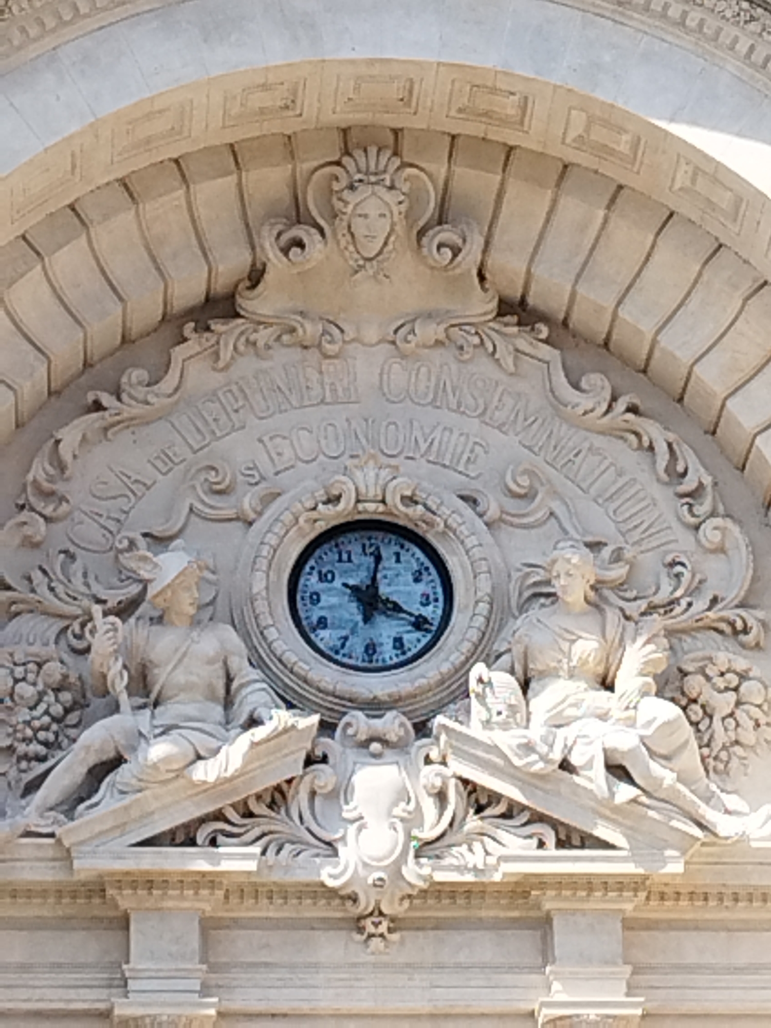 The image shows a close-up of an ornate architectural facade, likely a building in Bucharest, Romania. A prominent element is a large clock face, centrally positioned within elaborate stone carvings. The clock has a light blue face and black hands, indicating a time around 5:27. Flanking the clock are two neoclassical sculptures. The sculpture on the left depicts a male figure wearing a helmet and holding a staff, seated on a decorative ledge. The sculpture on the right shows a female figure, also seated, draped in classical attire. Both sculptures are rendered in light-colored stone. Above the clock and sculptures, curved stonework features carved lettering that reads "CASA DE DEPUNERI s ECONOMIE CONSEMNATIONI." The surrounding architecture is intricately detailed with relief carvings, including vegetal motifs and facial ornaments. The overall impression is one of historical grandeur and artistic craftsmanship.