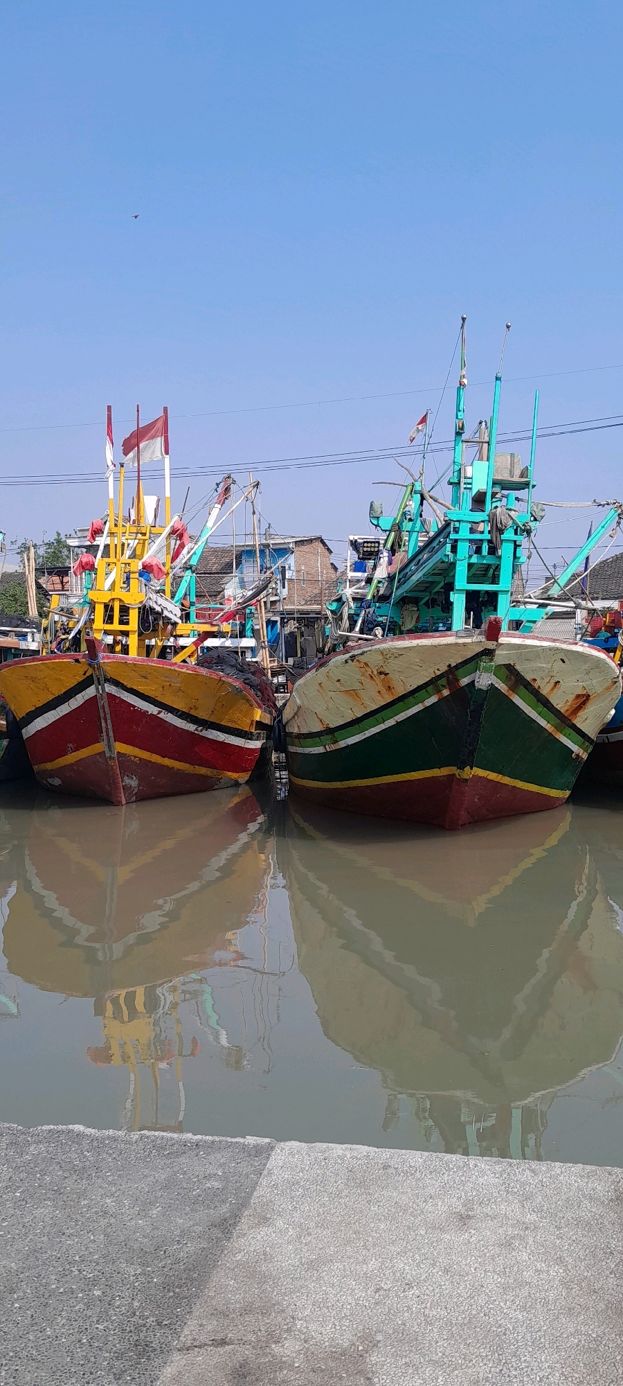 The image depicts a vibrant scene of several colorful fishing boats docked side-by-side in the city of Pasuruan, Indonesia. The boats are painted in bold red, yellow, and green hues, with white accents and some exhibiting signs of wear and tear, suggesting active use. Prominently displayed on the boats are Indonesian flags, indicating local ownership and national pride. The boats are reflected in the calm, murky water, creating a mirrored effect that adds depth to the composition. In the background, a cluster of buildings with tiled roofs and various electrical wires suggests an urban or semi-urban coastal environment. The sky is a clear, bright blue, indicative of fair weather. The foreground shows a textured gray surface, possibly a concrete dock or walkway. The overall atmosphere is one of a bustling fishing port, with the boats awaiting their next journey out to sea.
