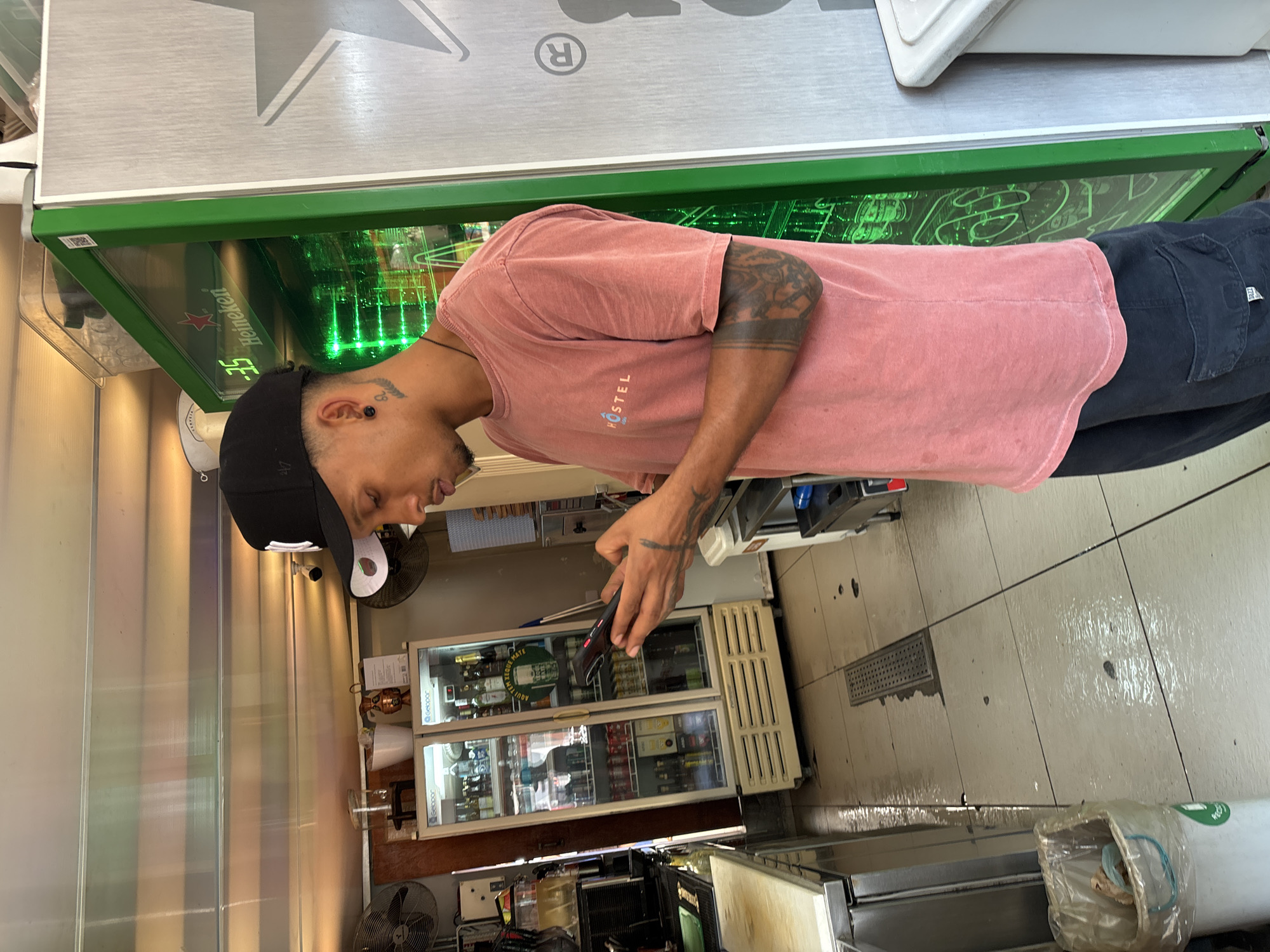 A man stands in what appears to be a commercial establishment, possibly a store or small business, in São Paulo, Brazil. He is wearing a pink t-shirt, dark-colored pants, and a black baseball cap, looking at his phone. The man has visible tattoos on his arms and head. Behind him are refrigerators stocked with beverages. In the background, there is a counter, flooring and some equipment. Overhead is a beverage cooler. The floor appears to be tiled and somewhat wet.