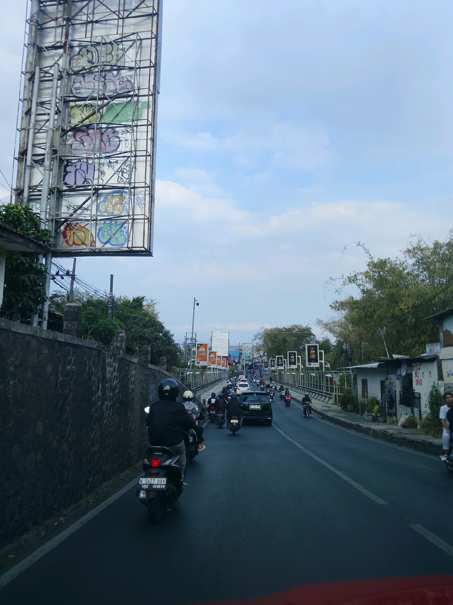 The image captures a busy street scene in Malang, Indonesia, likely on Jalan Soekarno Hatta, known for its traffic and billboards. In the foreground, a person on a black motorcycle with license plate "N 5627 BAX" is riding in the left lane of a multi-lane road. The motorcycle is facing away from the camera. Behind this motorcycle, a stream of other motorcycles and a few cars are visible, indicating moderate traffic congestion.
To the left of the road, a textured stone wall runs alongside the street, topped with greenery. Above the wall, a large, imposing billboard structure dominates the upper left portion of the image. This billboard is covered in various graffiti tags in multiple colors, suggesting an urban, perhaps slightly edgy, environment. Further down the road, more billboards are visible, some displaying advertisements.
On the right side of the road, buildings and trees line the street. Some of these buildings appear to be simple structures with roofs, and one has graffiti on its wall. The sky above is partly cloudy, with a light blue hue. The overall impression is of a typical day in a bustling Indonesian city, with a mix of modern infrastructure, urban art, and everyday life unfolding on the road.