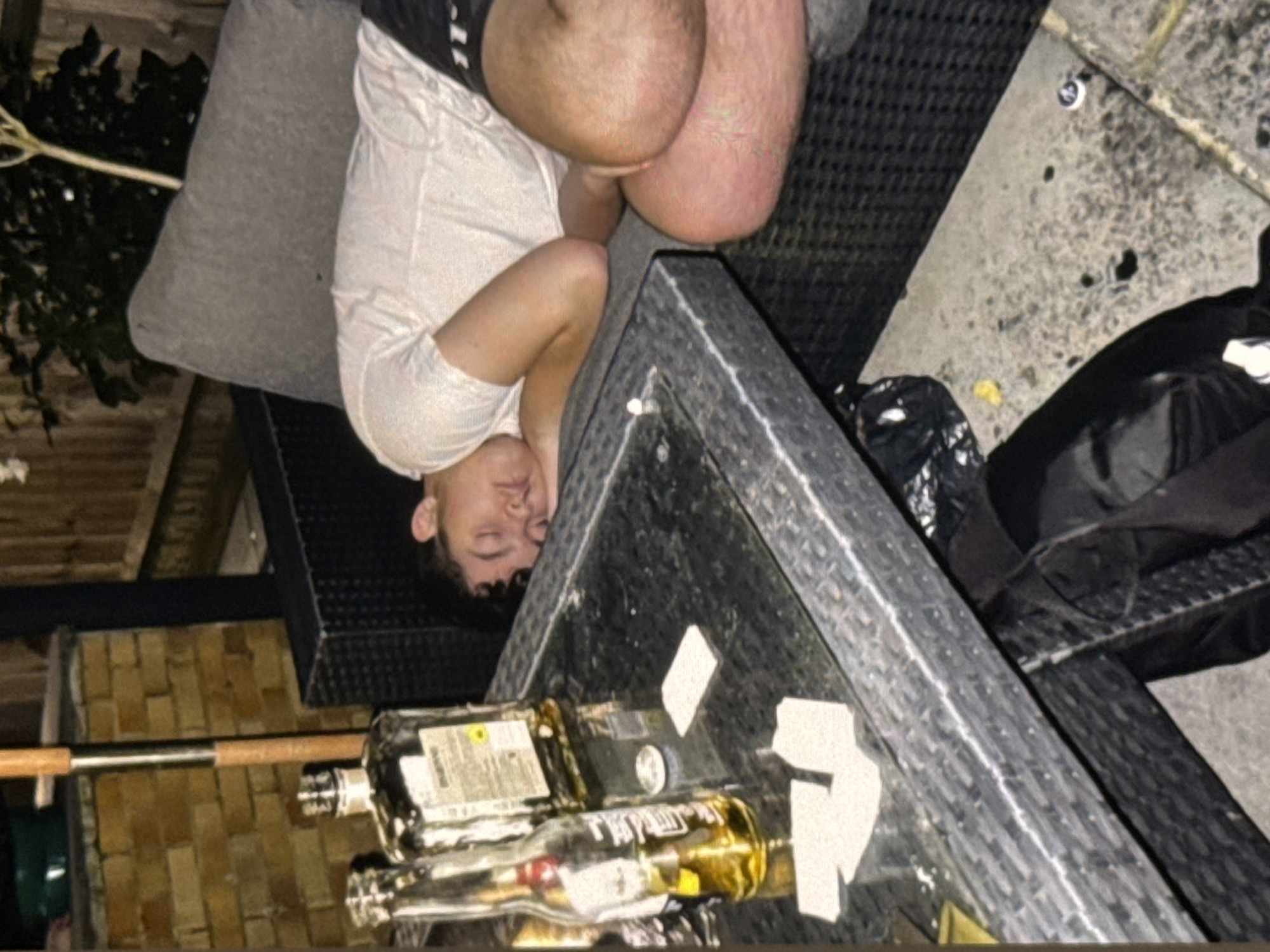 A young man with dark, curly hair is asleep on a grey outdoor sofa. He is wearing a white t-shirt and dark shorts. His head is resting on his arm. To his left is a dark grey, wicker-style outdoor table. On the table are two bottles, one clear with yellow liquid, and another, darker bottle. There are also several white rectangular objects on the table, possibly business cards or coasters. The scene appears to be an outdoor patio or garden area. The lighting suggests it is evening or night. The environment shows a brick wall behind the sofa and a wooden fence in the background. The ground is paved. There are also some black bags on the ground near the table. This image does not contain any discernible elements to indicate it is located in Yerevan, Armenia.