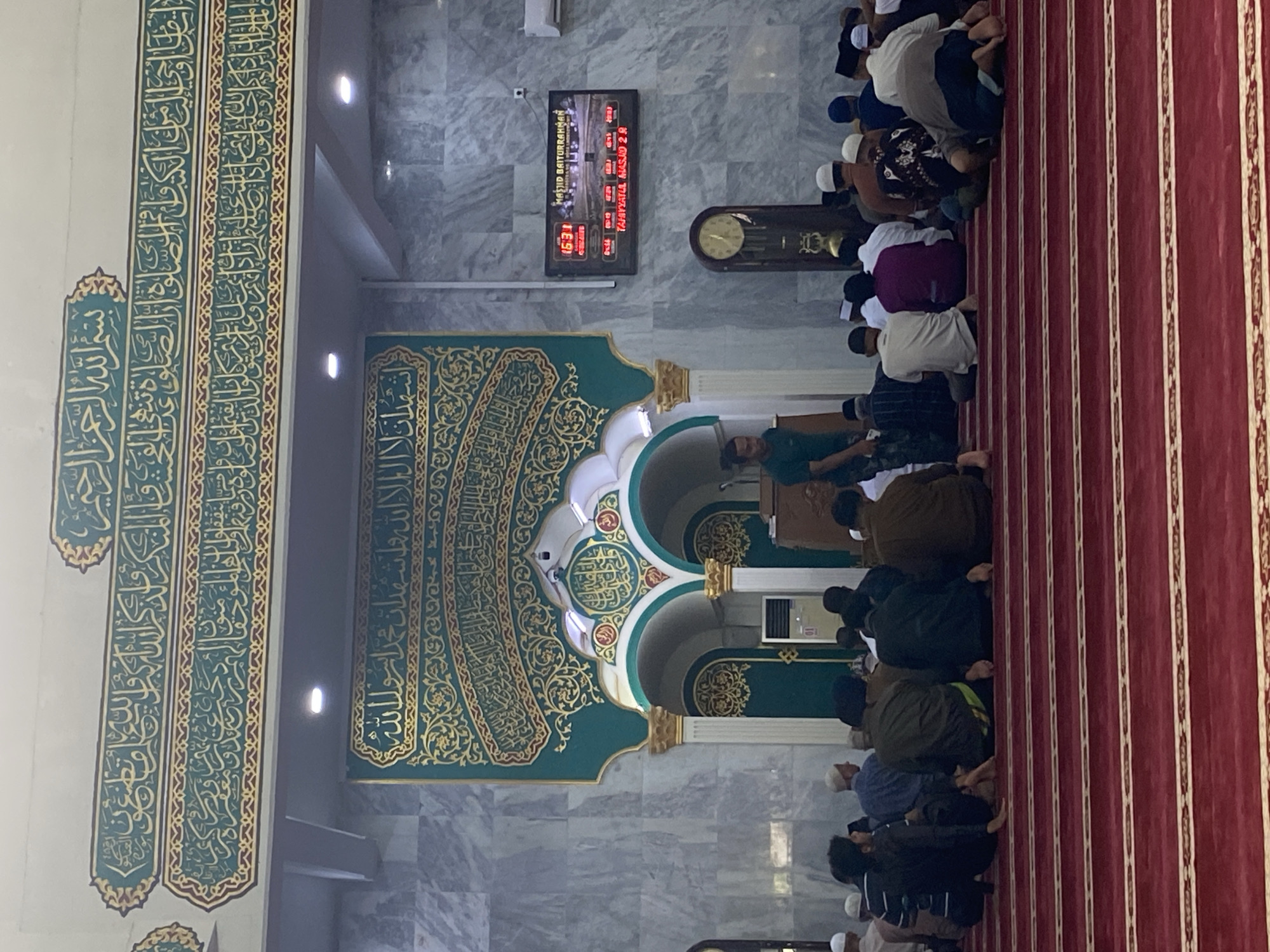 The image depicts the interior of a mosque, likely Masjid Baiturrahman in Lhokseumawe, Indonesia, based on the digital clock display which shows "MASJID BAITURRAHMAN". Several men are seated on a red carpet with gold patterned lines, facing towards the front of the mosque, indicating they are likely engaged in prayer or awaiting prayer. Some are in a sitting position, while others are in a prostrated position, common during Islamic prayers.
The mosque features ornate green and gold Arabic calligraphy on its walls and above the arched doorways. There's a large digital display showing prayer times and the mosque's name. A grandfather clock is visible on the right wall. An air conditioning unit is mounted on the wall near the entrance. The walls are clad in a marble-like material. A man in a green shirt appears to be standing near the prayer area, possibly a mosque official or a participant. The overall atmosphere is one of reverence and communal worship.