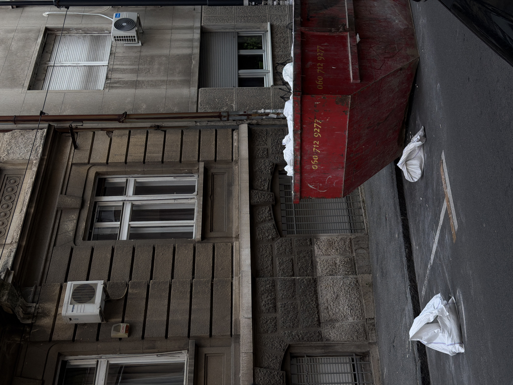 The image shows a building facade with several windows and two air conditioning units. A large, red dumpster partially obscures the right side of the building and extends onto the street. There are two white bags on the street near the dumpster. The building appears to be located in a street. The style of architecture and presence of the dumpster suggest the area may be undergoing construction or renovation. The phone number "060 712 9277" is visible on the side of the dumpster. The image depicts a scene in a busy urban area, potentially located in Vracar Urban Municipality, Serbia. The buildings are constructed out of rock. The photo is taken during daytime.