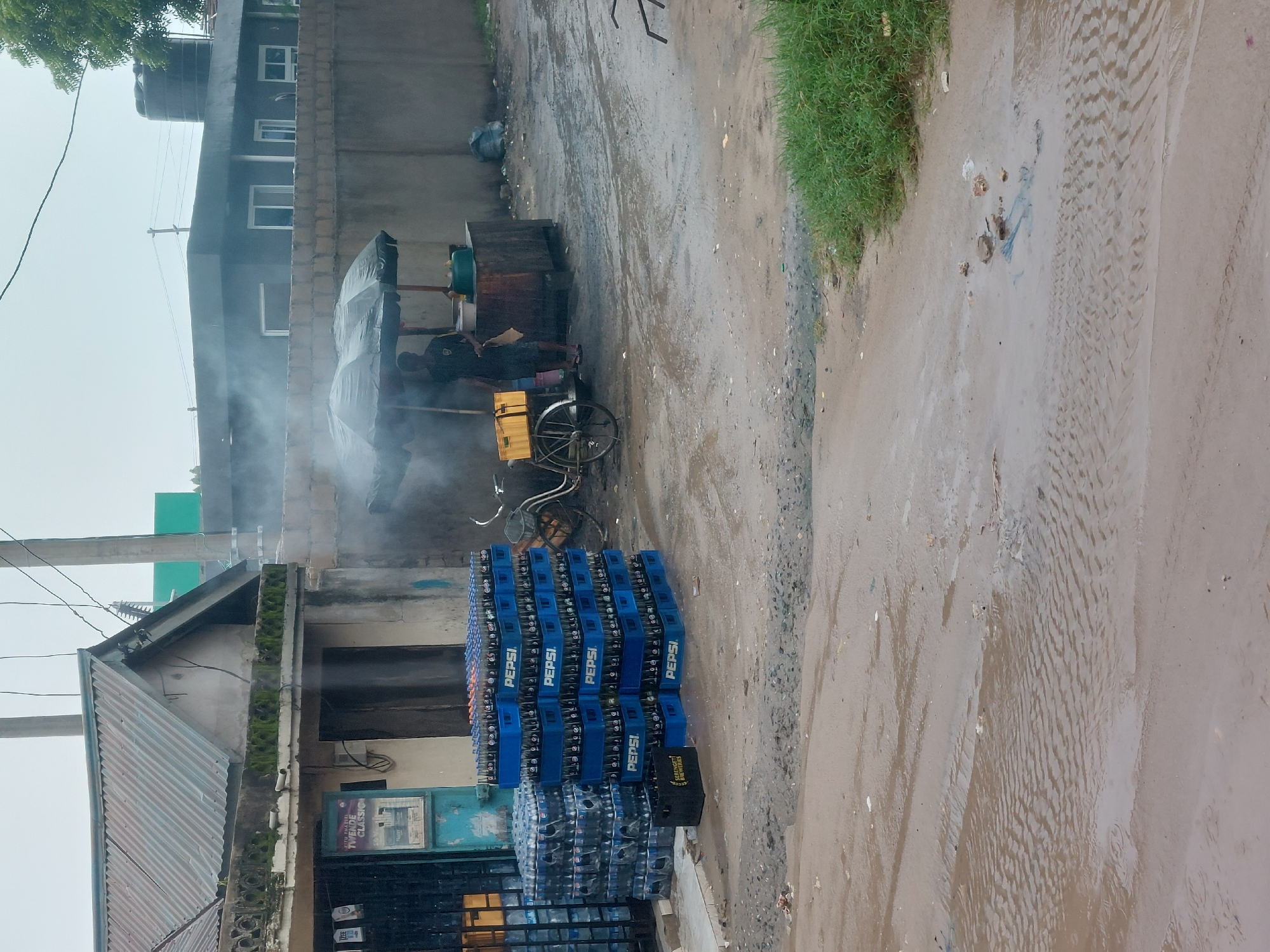 The image depicts a street scene in what appears to be a residential area. The street is partially flooded with muddy water. A stack of Pepsi and water cases sits near a building with a corrugated metal roof. A mobile food cart with steam rising from it is positioned further down the street, with at least one person standing near it. A bicycle with yellow crates on the back is parked adjacent to the cart. In the background are residential buildings. I don't have enough information to determine that this scene is located in Dar es-Salaam, Tanzania.