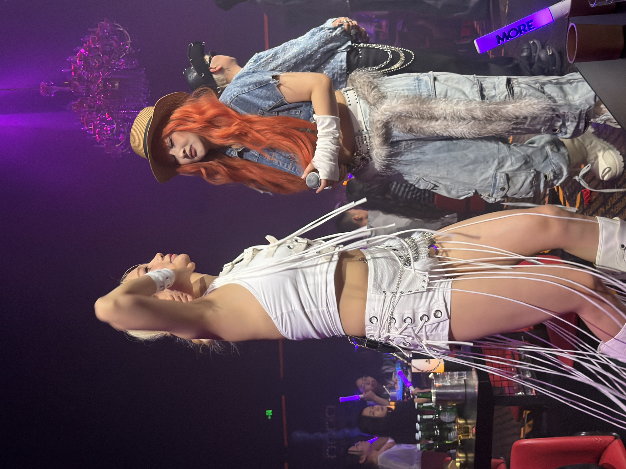 In a nightclub in Jiangbeichen Street, Chongqing, China, two young women perform on a dimly lit stage under purple spotlights. The woman on the left, with blonde hair, wears a white halter top and white shorts adorned with numerous white tassels and silver chains. She is posing with her arm raised. The woman on the right, with bright orange hair, wears a denim jacket over a white top, light blue baggy cargo pants, a straw hat, and white wrist warmers. She holds a microphone and stands with her eyes closed, seemingly singing. In the background, other patrons are visible at tables, with bottles of beer and a chandelier hanging overhead. A purple glow stick with the word "MORE" written on it is held by an unseen person in the foreground.