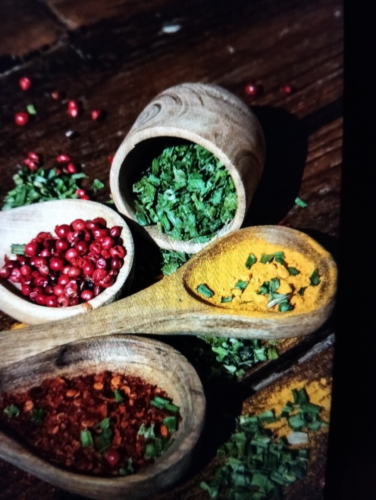 The image depicts a rustic arrangement of spices on a wooden surface. In the foreground, a wooden spoon contains red chili flakes and what appear to be chopped green herbs. Adjacent to it, another wooden spoon holds yellow turmeric powder, also mixed with green herbs. To the left, a small wooden bowl is filled with vibrant red peppercorns. Behind these, a larger wooden bowl is overflowing with dried green herbs. Scattered across the wooden surface are a few stray red peppercorns and green herb pieces. The lighting creates strong shadows, suggesting an indoor setting with directional light. There is no discernible information within the image to indicate a specific location in Payagaboh, Indonesia, or any human presence.