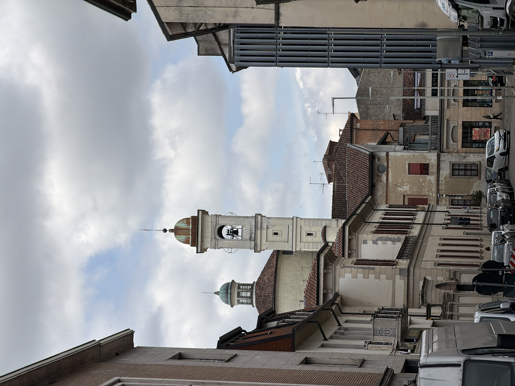 This image depicts a daytime urban street scene in Vigevano, Italy, viewed from a slightly elevated or angled perspective, looking down a street or alley. The original image is rotated, so the description assumes an upright orientation.
The scene is dominated by a tall, light-colored bell tower (campanile) on the left side, featuring a distinctive oxidized green dome and a spire. A bell is visible within its belfry. To the left of the bell tower, a smaller, similar green-domed structure is also present, likely part of the same religious complex or historic building. The surrounding buildings showcase traditional European architecture, with some featuring terracotta tiled roofs and aged facades, while others present more modern or industrial elements, such as a gray horizontally-slatted structure on the right.
The street itself is lined with parked vehicles, including several vans, one white and one silver, along with other cars, particularly on the right side. Street lamps and various signs are visible. On the right, a building facade displays the partial text "FARMA," suggesting a pharmacy (Farmacia). A person is seen walking in the distance on the right side of the street. Notable street signs include a "P" for parking and a red circular prohibitory sign with an arrow.
The sky is partly cloudy, with patches of brighter blue visible amidst a generally overcast backdrop, indicating a moderate to bright day. The overall impression is that of a typical, active street in a historic European town.