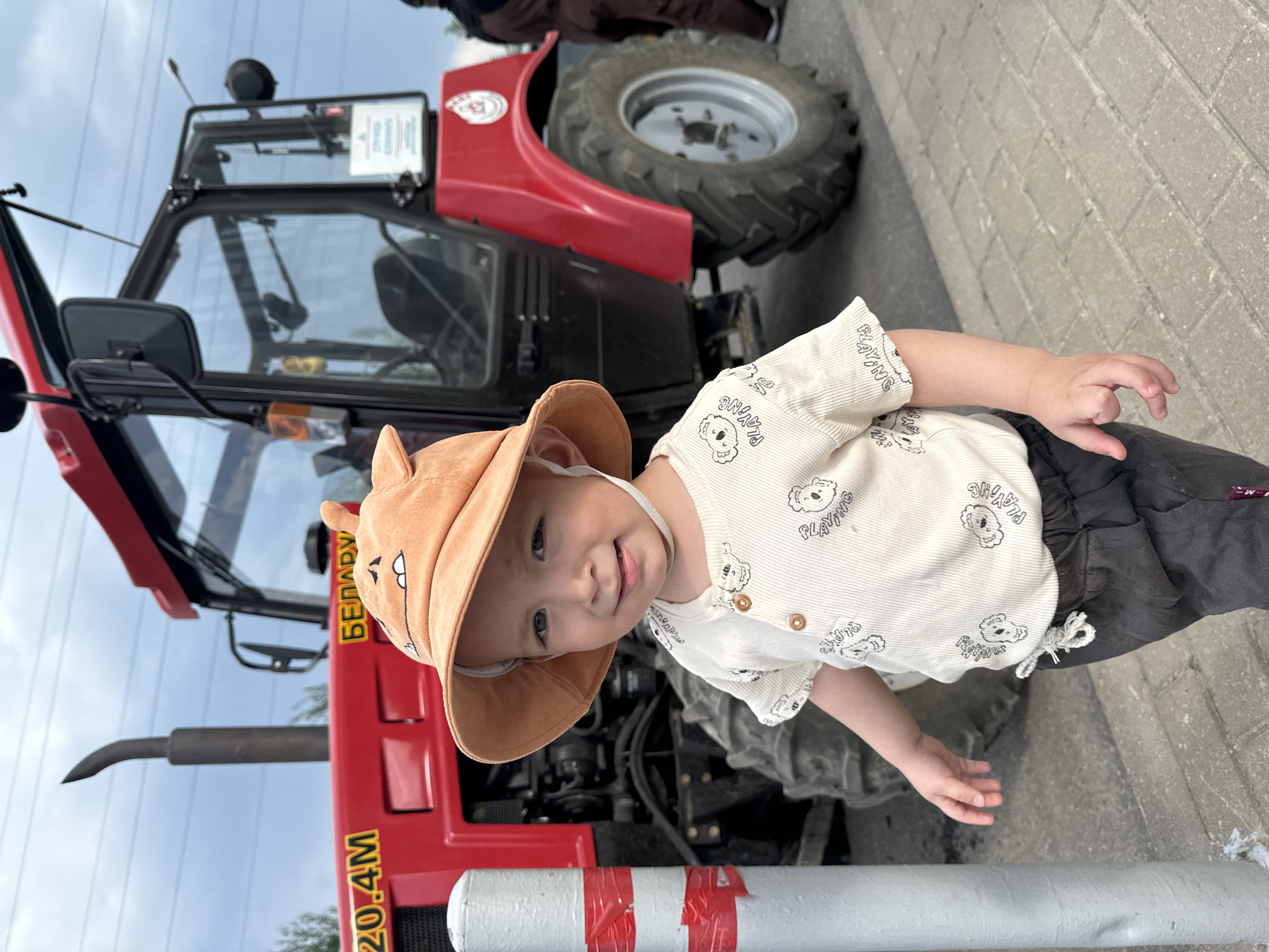 A child stands in front of a red and black Belarus tractor model 820.4M in Odintsovo, Russia. The child wears a tan sun hat with ears and a white shirt with bear designs. The tractor is parked on a paved surface. Overhead are electrical lines against a cloudy sky.
