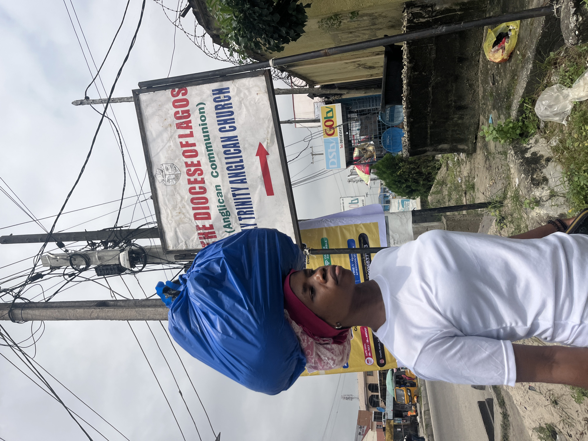In Ado, Nigeria, a woman walks along a street, carrying a large blue plastic bag filled with items balanced on her head. She wears a white t-shirt and a pink headscarf. Behind her, a sign reads "THE DIOCESE OF LAGOS (Anglican Communion) HOLY TRINITY ANGLICAN CHURCH" with a red arrow pointing to the right. The scene is characterized by numerous overhead electrical wires, a utility pole, and a concrete wall with barbed wire on top. To the left of the woman, a yellow motorized tricycle (Keke Napep) is visible on the street, and further down the road, buildings and shops line the street. The ground is unpaved and covered with scattered debris, including plastic bags and vegetation. The sky is overcast, suggesting a cloudy day.